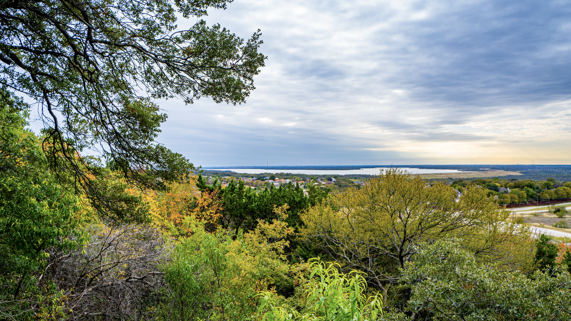 View from a hillside with green and autumn-yellow trees in the foreground; a distant town and lake lie on the horizon under a cloudy blue sky.