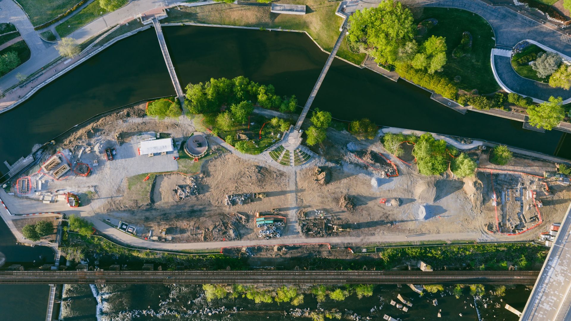 Aerial view of a river with pedestrian bridges, green trees along the banks, a circular plaza with a fountain, paths, and a construction site along the shoreline; railway tracks run along the bottom.