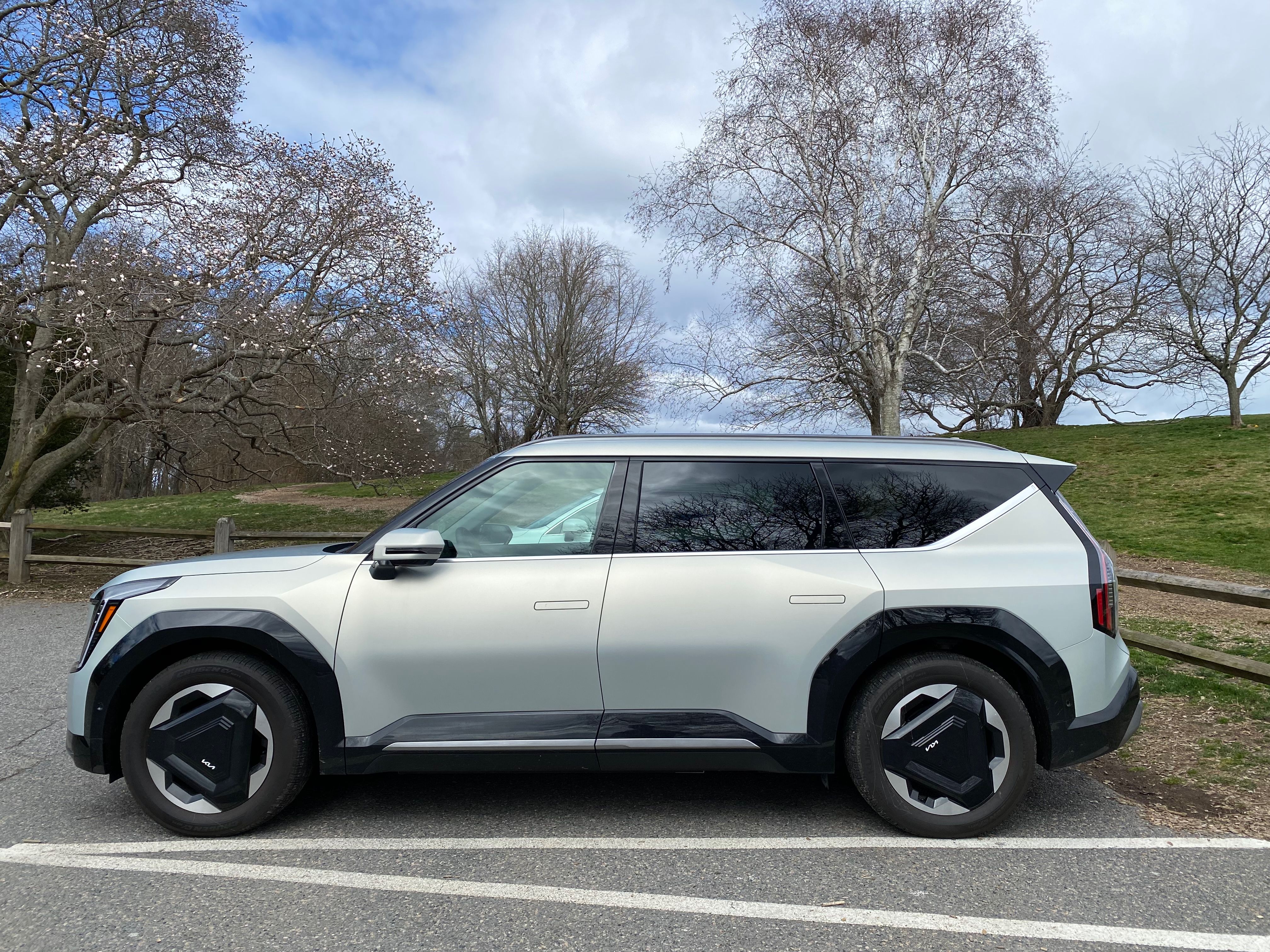 Side view of a silver Kia EV9 electric SUV parked on asphalt, with bare trees and grassy hill in the background under a partly cloudy sky.