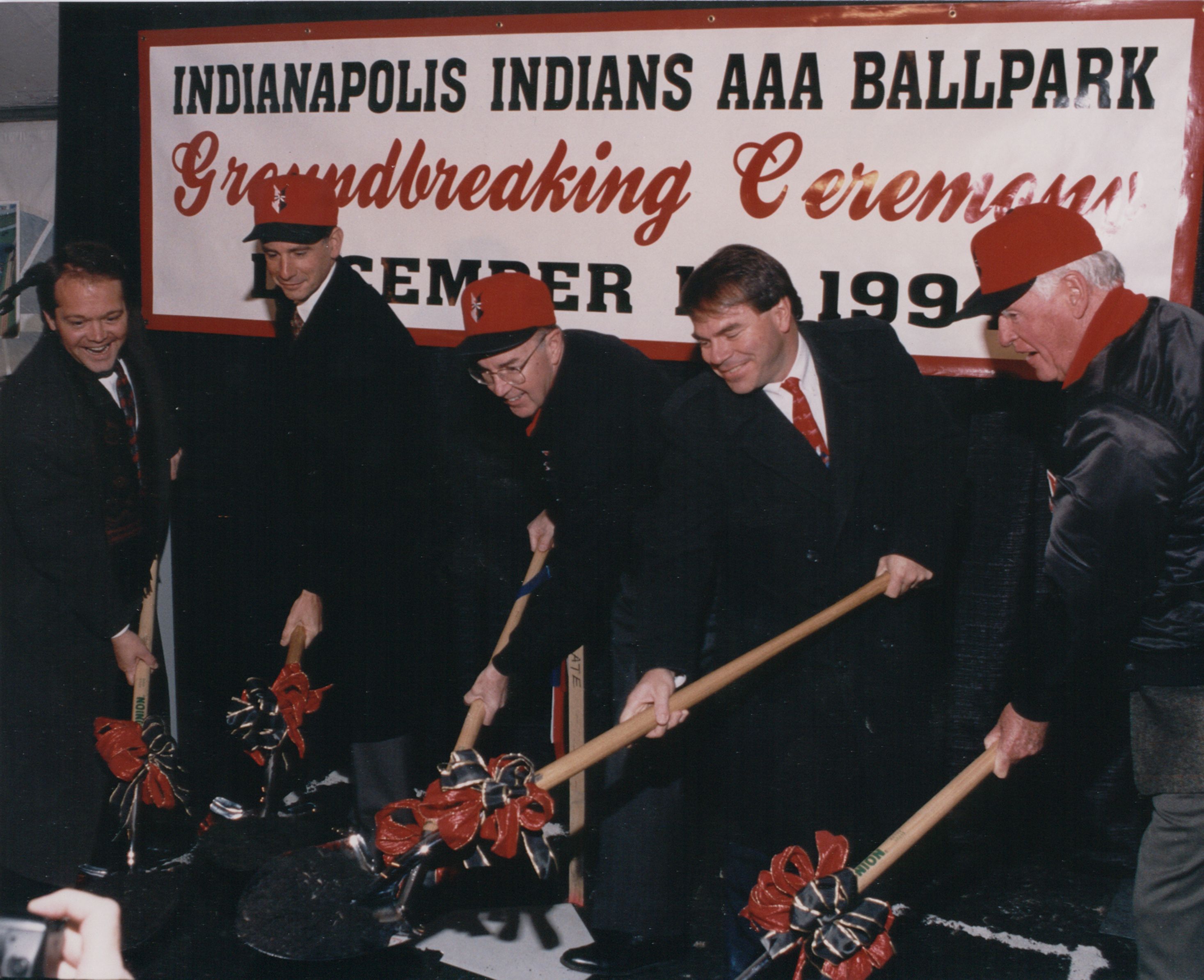 Five men in dark suits and red hats stand with shovels in front of a large banner that reads Indianapolis Indians AAA Ballpark and Groundbreaking Ceremony.