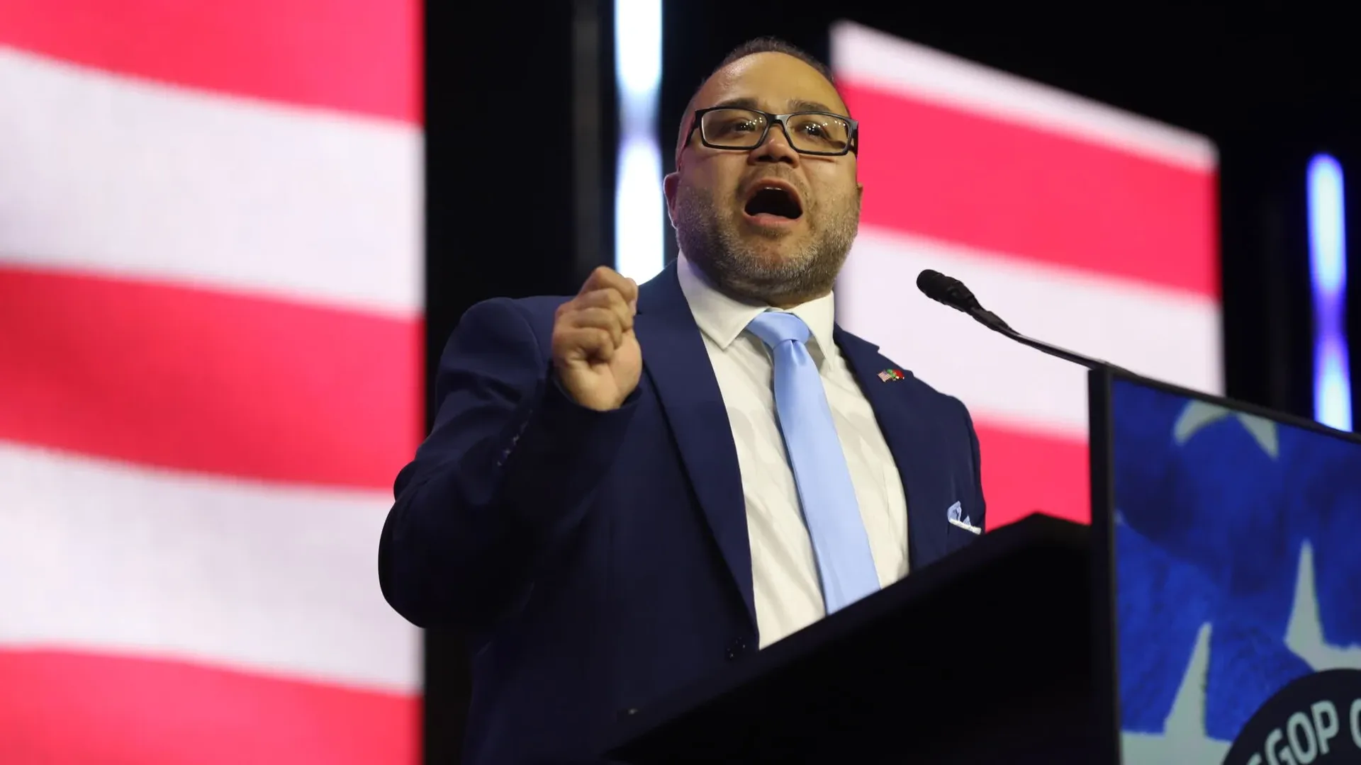 A man in a blue suit and light blue tie speaks at a podium, wearing glasses, mouth open as he delivers a speech; large red-and-white striped banners backdrop the stage.