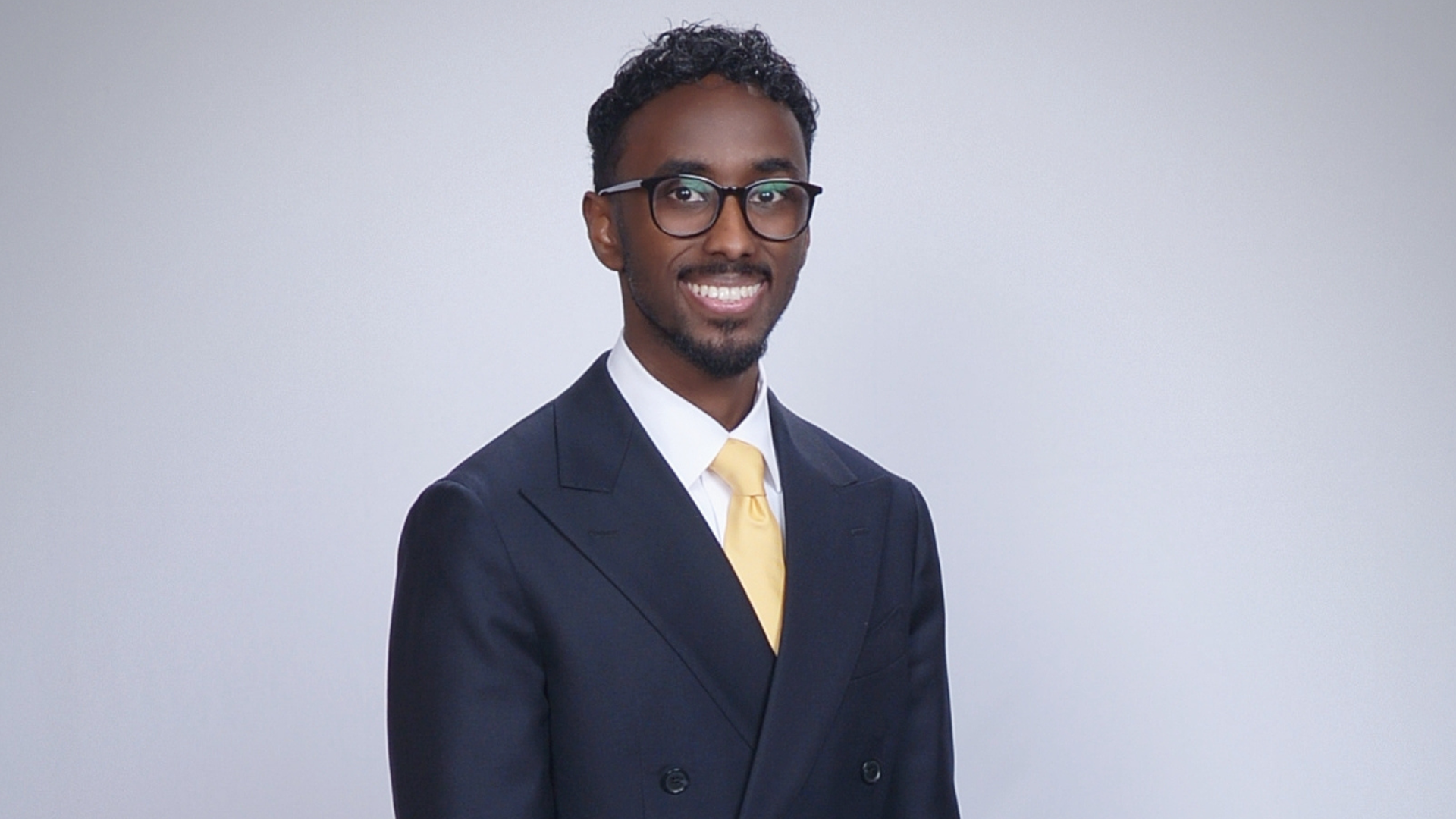 Smiling man in a dark double-breasted suit, white shirt, yellow tie, and glasses, posed against a plain light-gray backdrop.