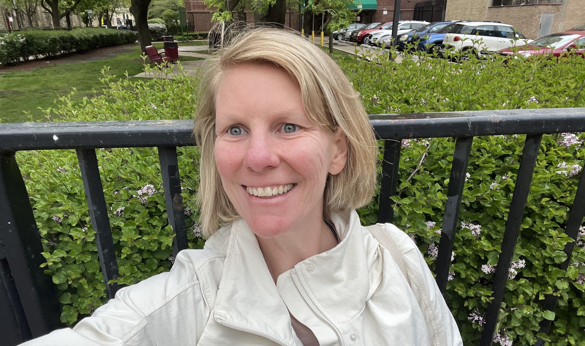Selfie of a Carrie Shepherd smiling outdoors, with a black metal railing in the foreground, green hedges and pink flowers behind, and parked cars and brick buildings in the background.