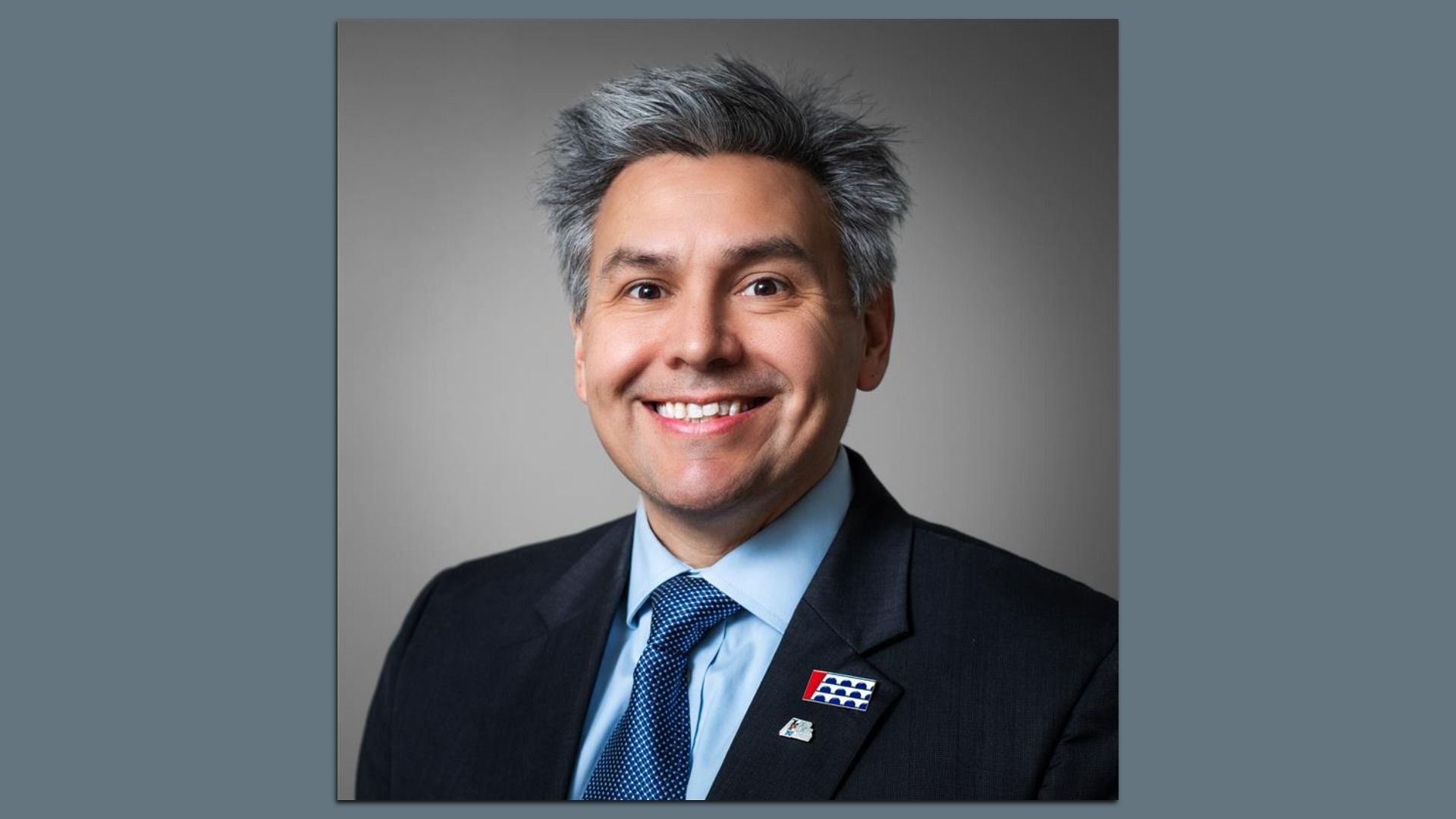 Smiling man with gray hair wearing a dark suit, blue shirt, and blue patterned tie, against a plain gray background, with two colorful lapel pins on his suit jacket.