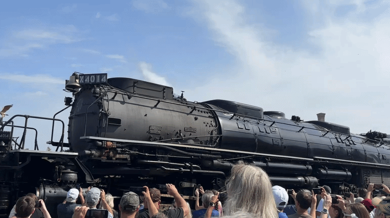 GIF shows panning view of a large black locomotive engine labeled No. 4014 blowing steam with a crowd gathered in the foreground watching and taking video on their phones.