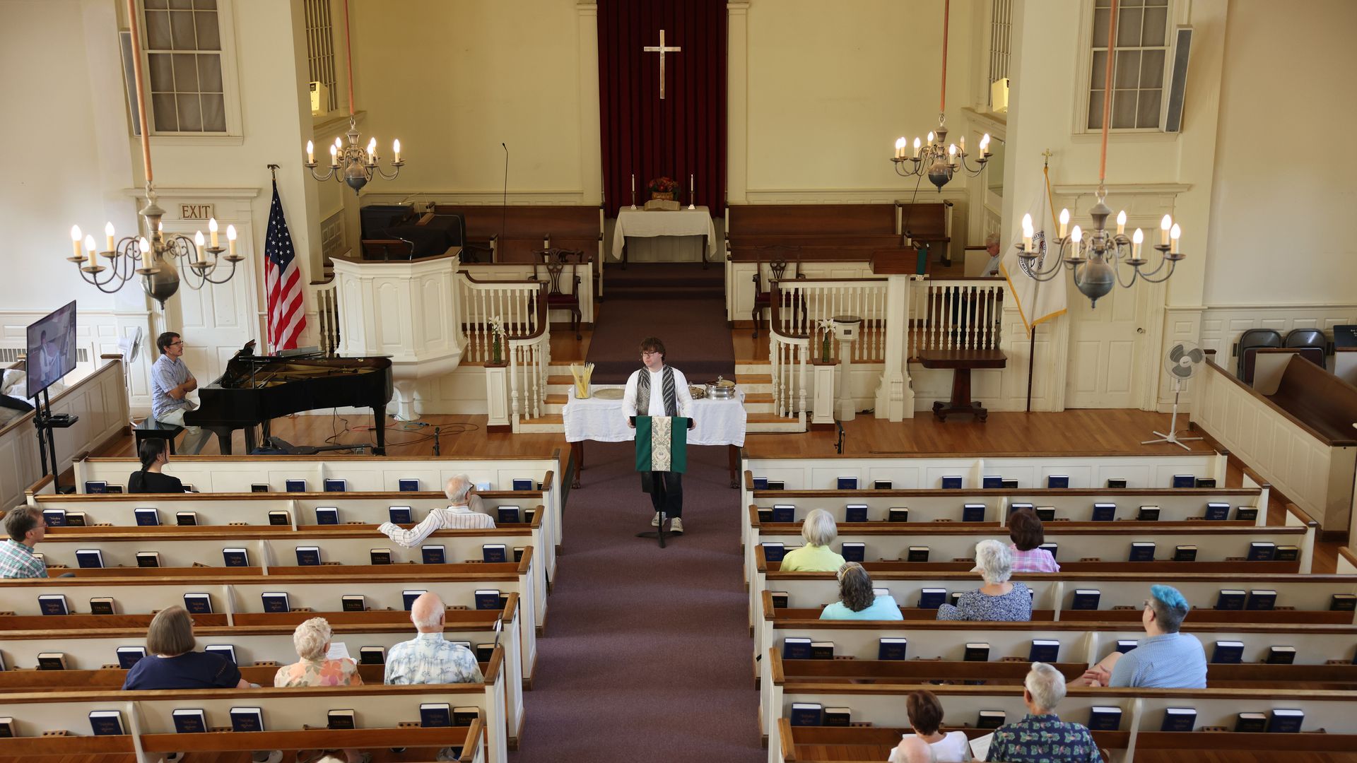 Inside a church with wooden pews and chandeliers, a minister in a white robe stands at the pulpit, addressing a small congregation seated sparsely throughout the pews.