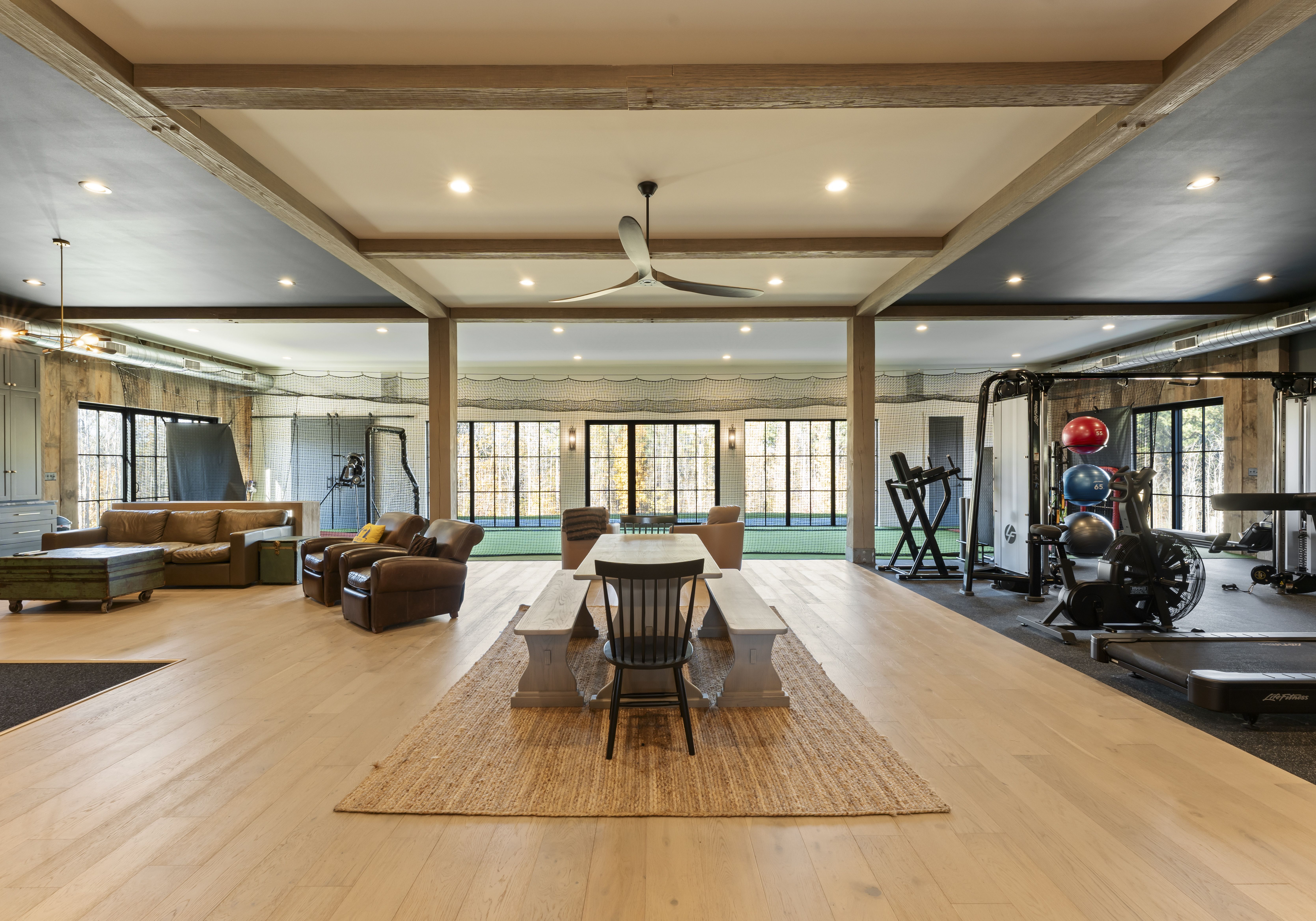 Spacious basement gym and lounge: a long white wooden table with benches sits on a woven rug, flanked by leather seating on the left and workout machines on the right; large windows in the back.