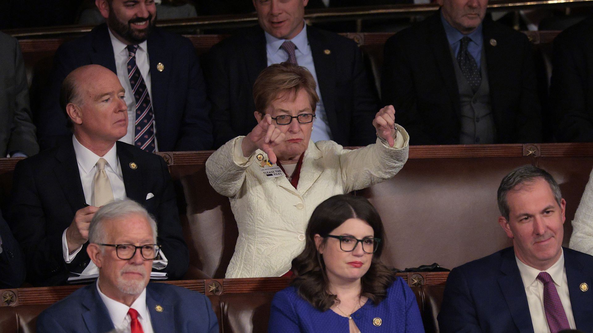 People in a formal audience seated in a theater balcony; a woman in a cream jacket at the center gestures with her hands, surrounded by men in dark suits and ties.