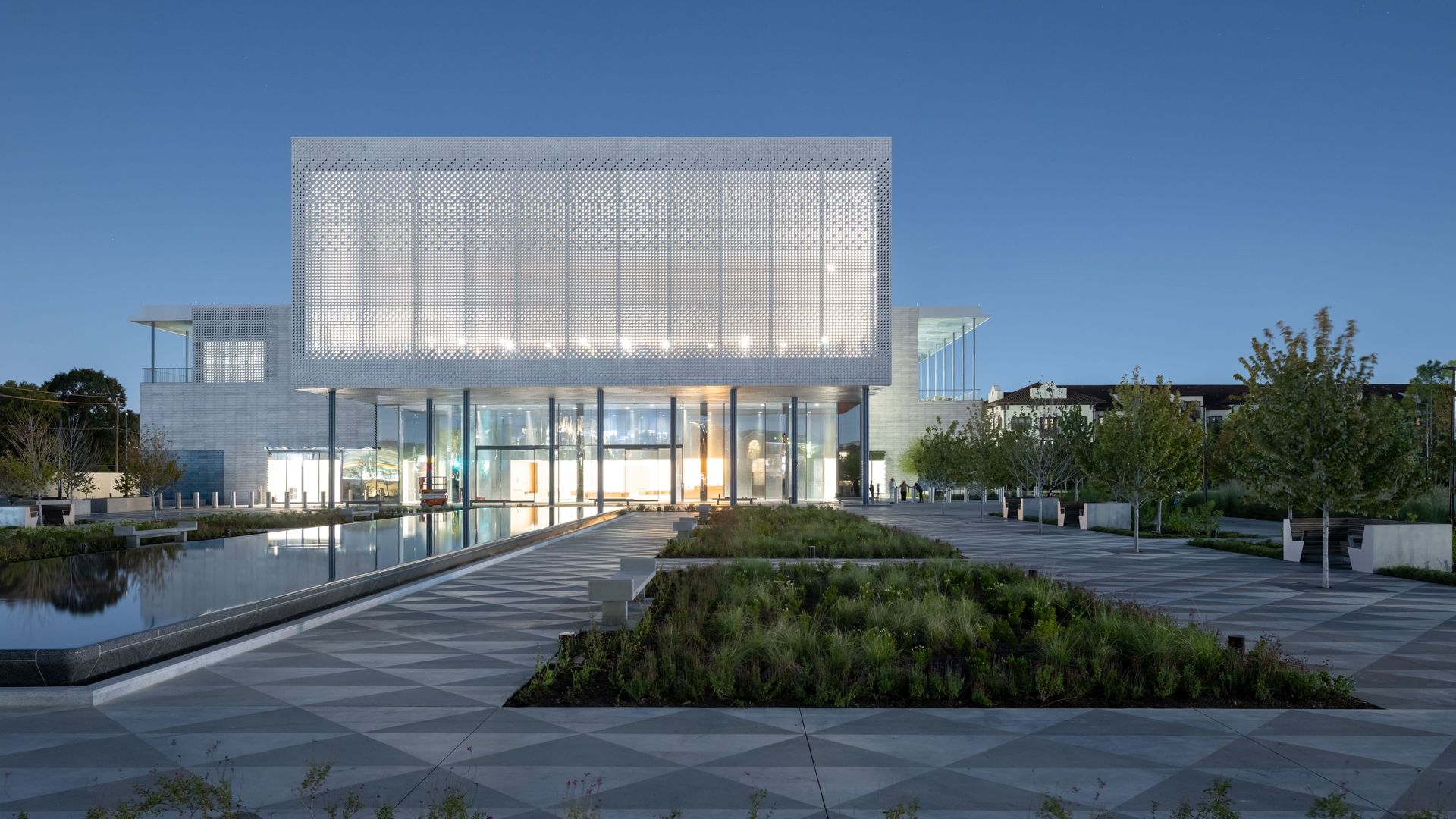 Modern building with illuminated perforated upper facade, glass entrance, reflective water feature, tiled walkway, and landscaped greenery under twilight sky.