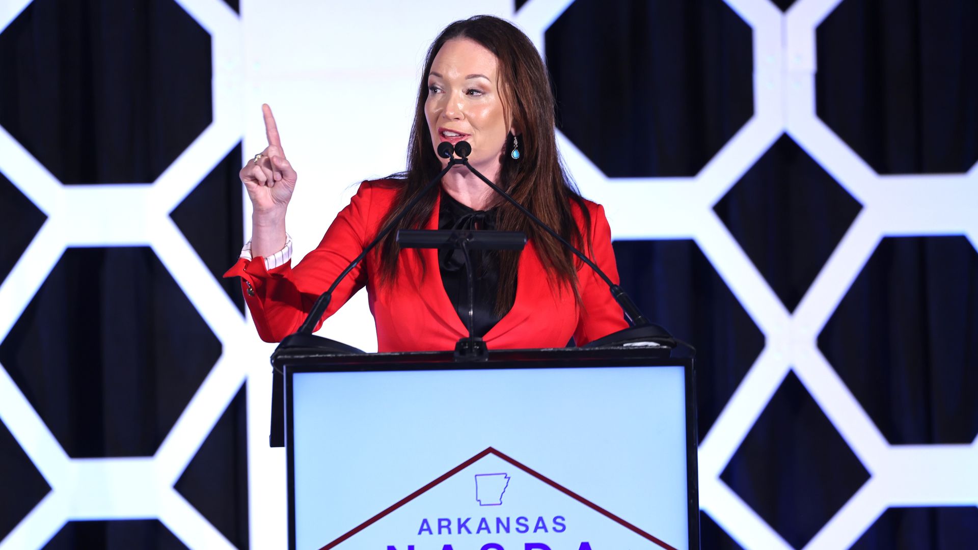 U.S. Agriculture Secretary Brooke Rollins in a red blazer is speaking at a podium with microphones. The podium sign reads "Arkansas NASDA 2025 Securing our Future through Agriculture" against a geometric white and black background.