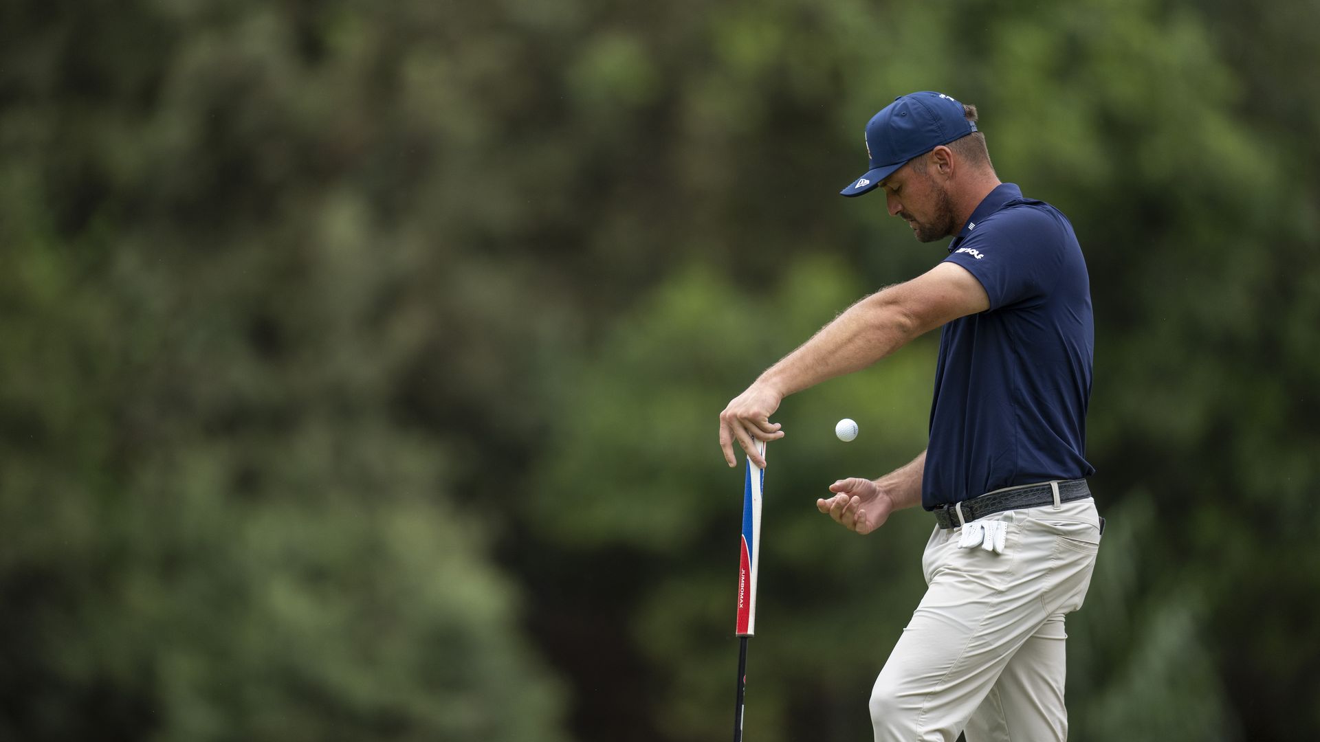 A golfer wearing a blue cap and navy shirt holds a club and tosses a white golf ball in the air with blurred trees in the background.