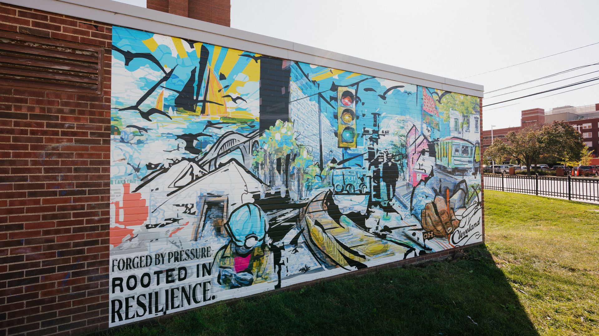Colorful mural on a brick wall shows a blue and yellow cityscape with birds and a traffic light; bold text reads "FORGED BY PRESSURE" and "RESILIENCE." A sunny day with grass and trees in the background.