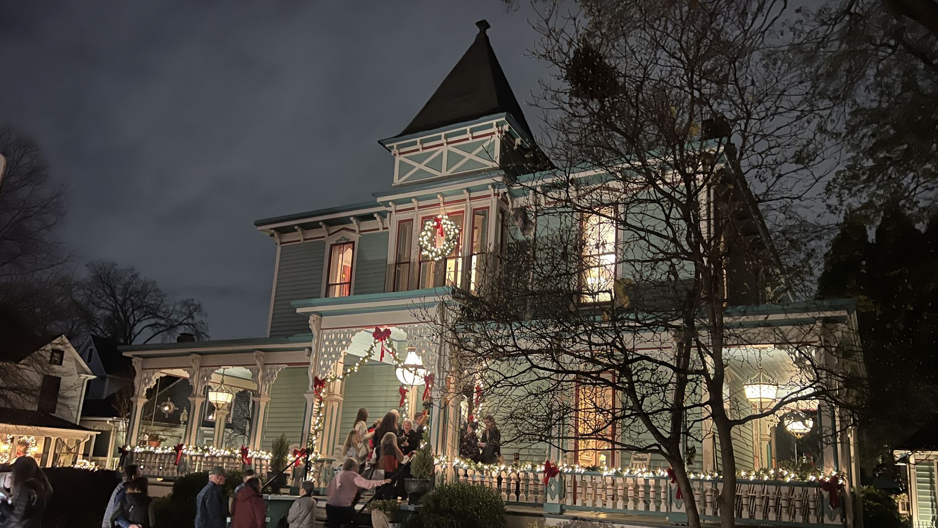 Victorian-style blue house decorated with white Christmas lights and red bows at night. A group of people is gathering and climbing the front porch steps under a lit wreath and garlands.