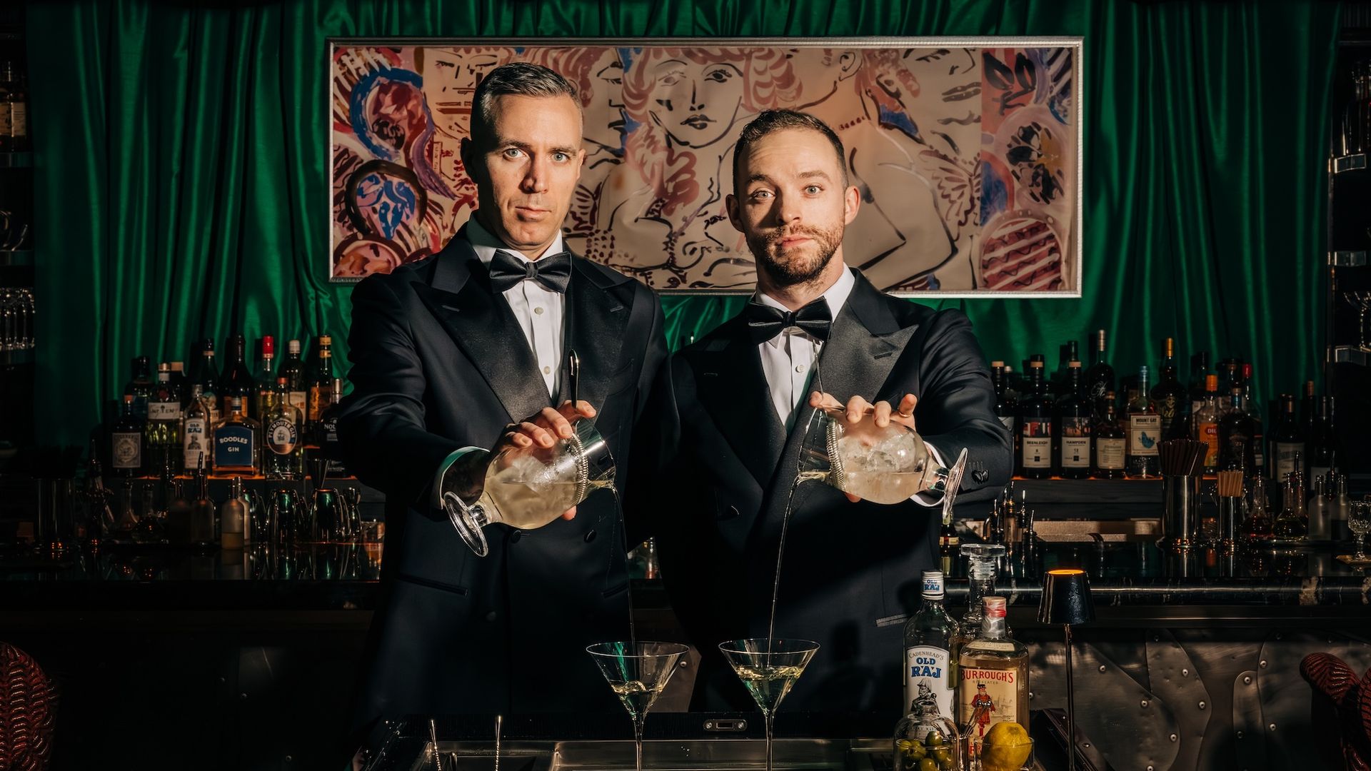 Two bartenders in black tuxedos pour cocktails from shakers behind a green velvet bar, with a colorful abstract painting and liquor bottles on the shelves; two martini glasses sit on the counter.