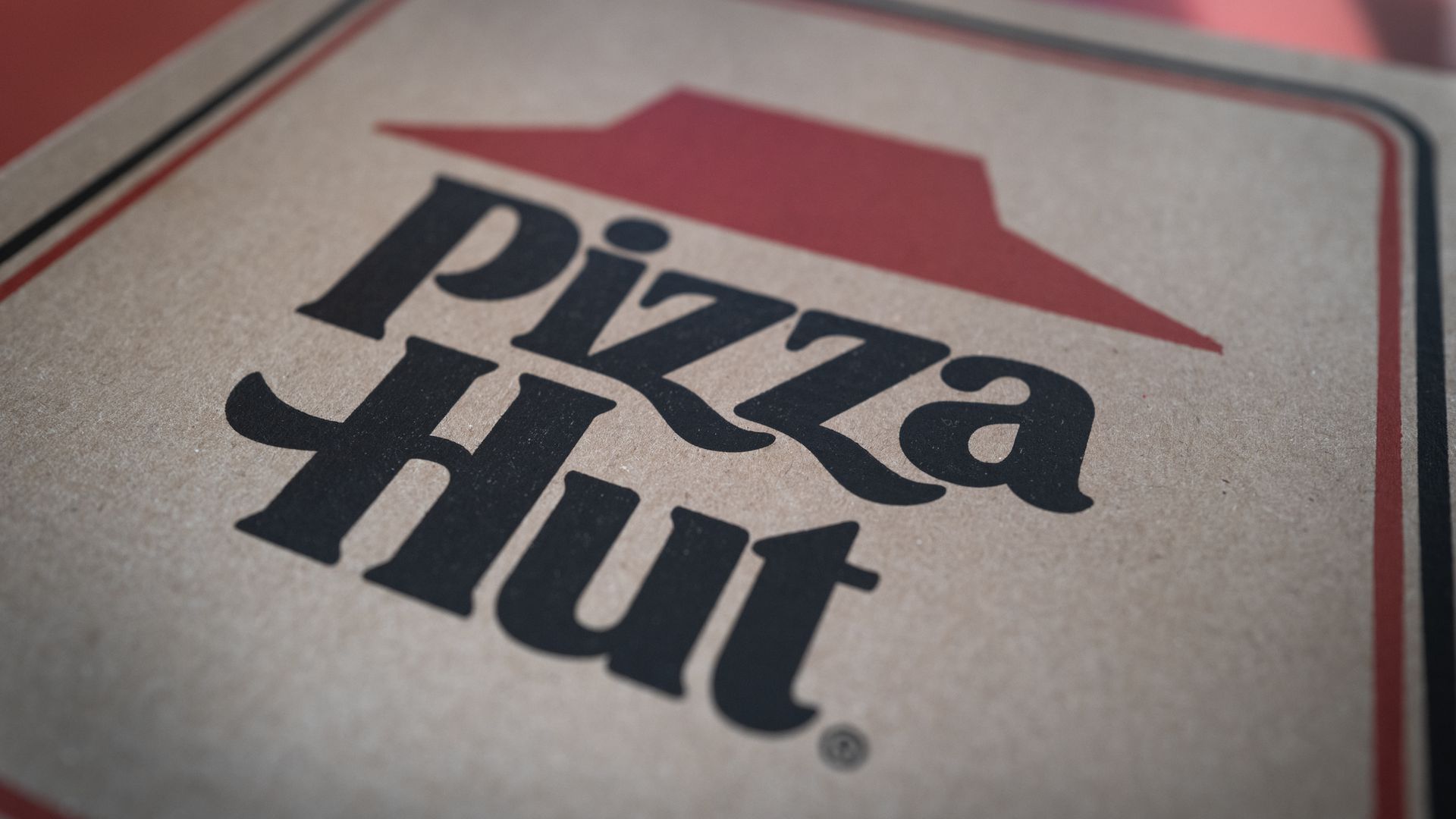 Close-up of a Pizza Hut logo on a cardboard pizza box, featuring a red roof above bold black lettering that reads Pizza Hut on a beige, textured surface.
