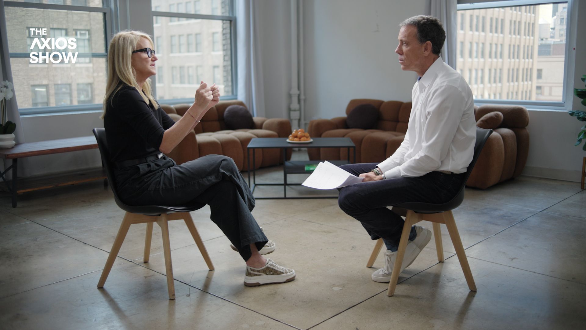 Robbins and VandeHei seated facing each other in a modern room with large windows and brown couches. Robbins gestures while VandeHei listens holding papers. "The Axios Show" logo on window.