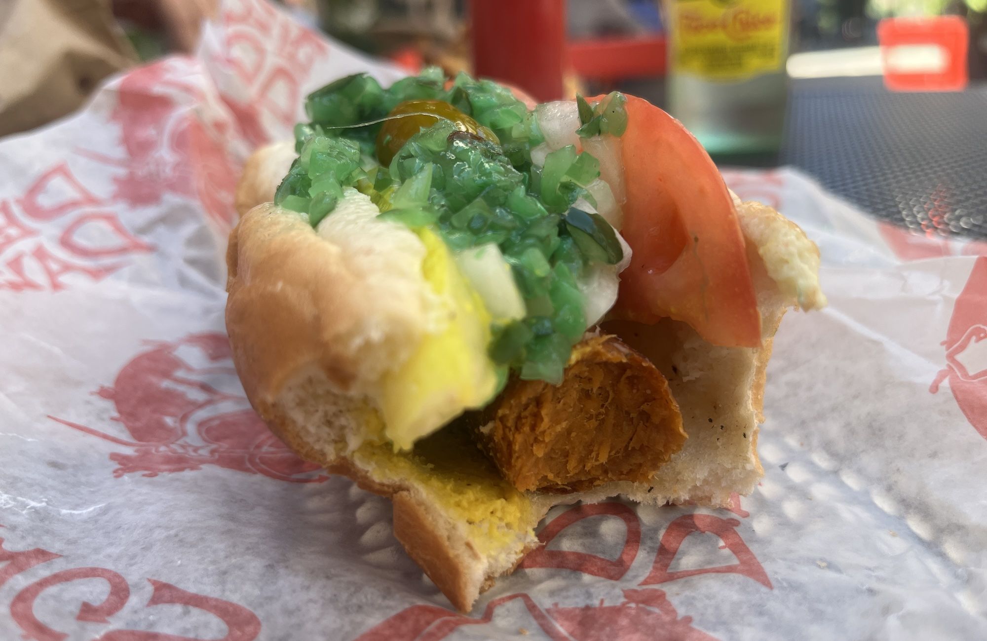 Close-up of a sandwich on a golden bun with yellow egg filling, white interior, bright green chopped relish, and a tomato slice; blurred outdoor food stand in the background.