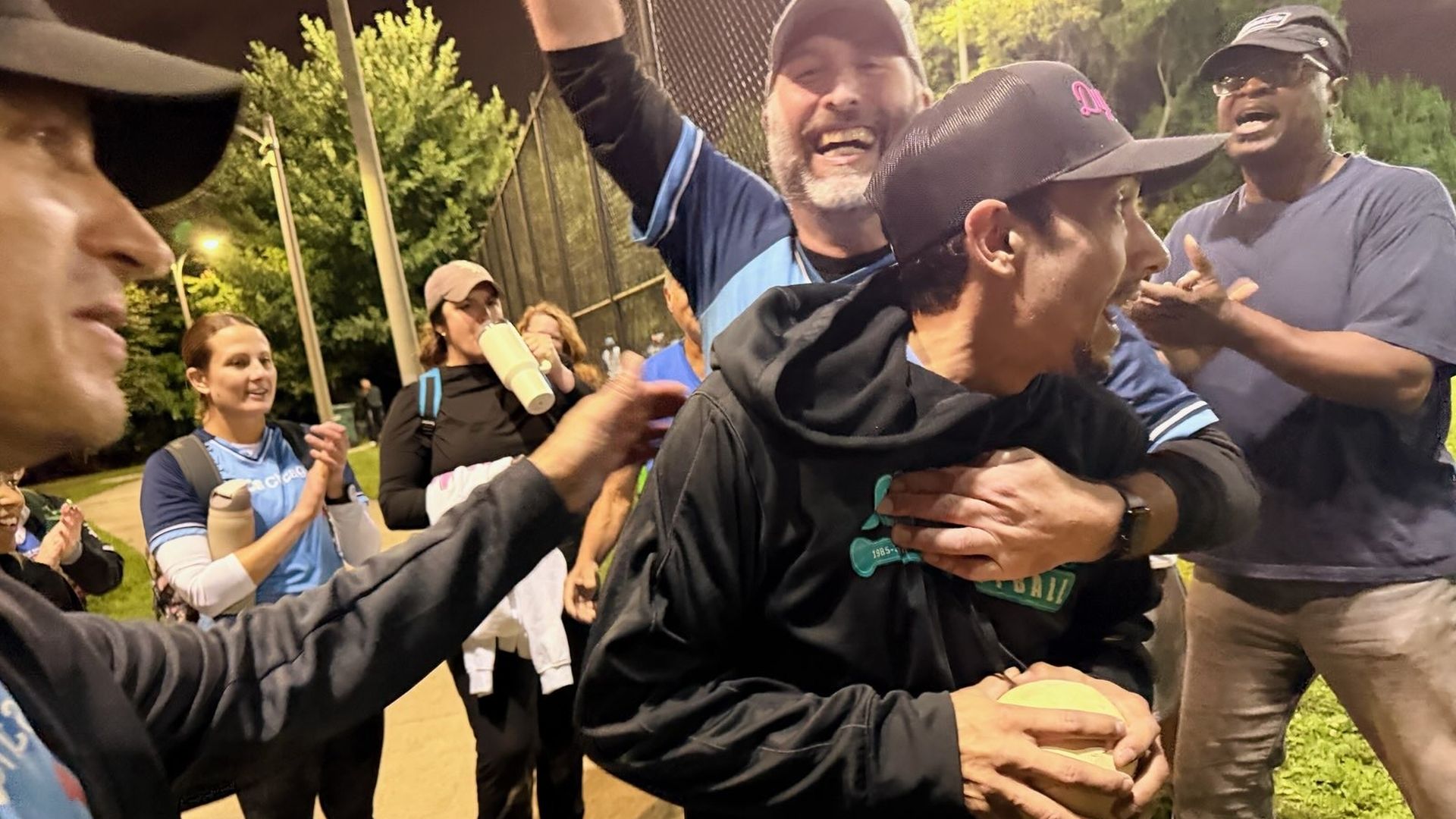 Nighttime group celebrating on a baseball field. A man in a black hoodie is hugged from behind by a smiling man in a blue shirt, while others cheer, clap, and watch; trees and fencing in the background.