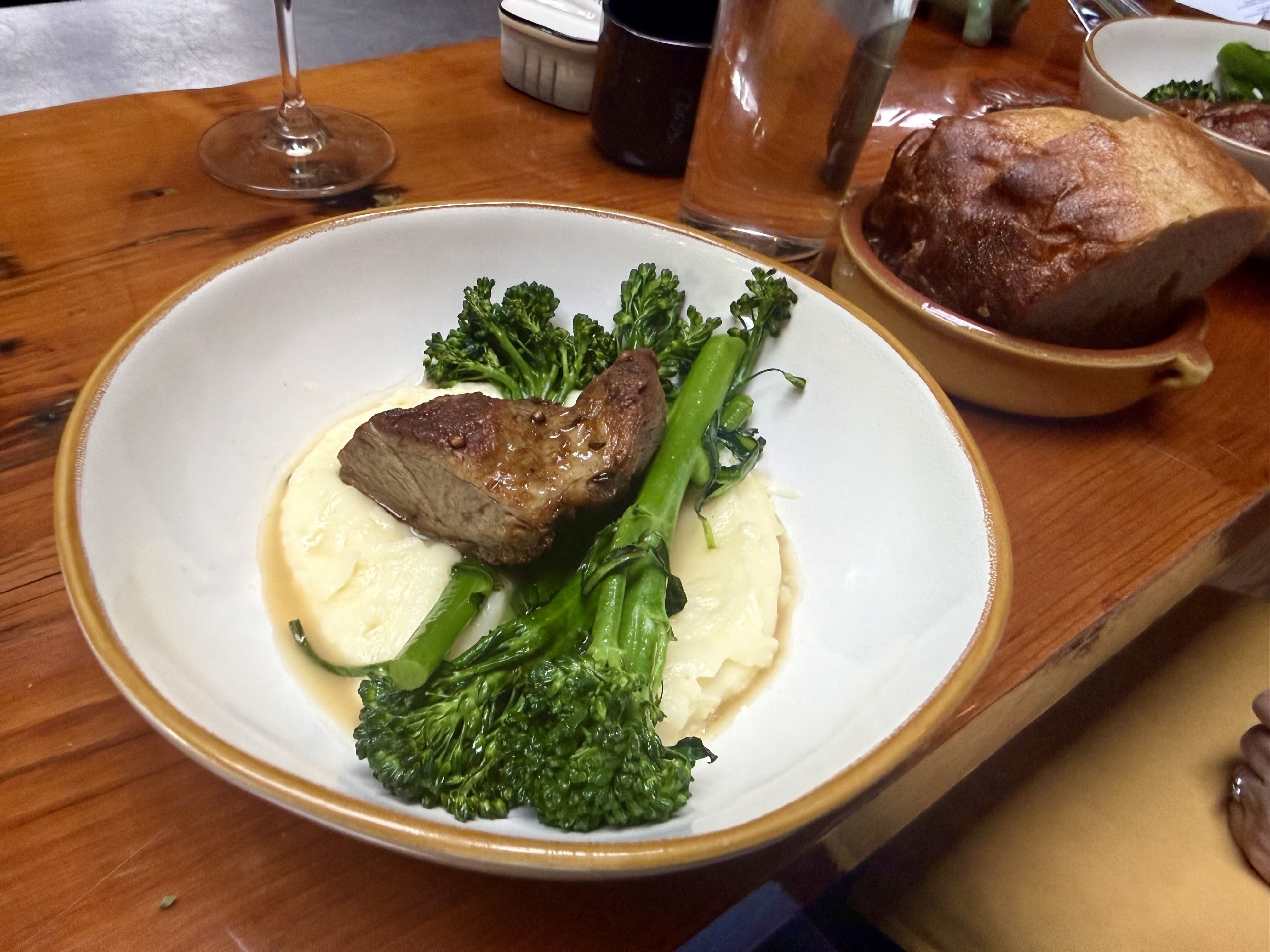 Rustic wooden table with a plate of browned lamb on creamy mashed potatoes, topped by green broccolini; a crusty loaf in a dish to the side; a wine glass and a water glass in the background.