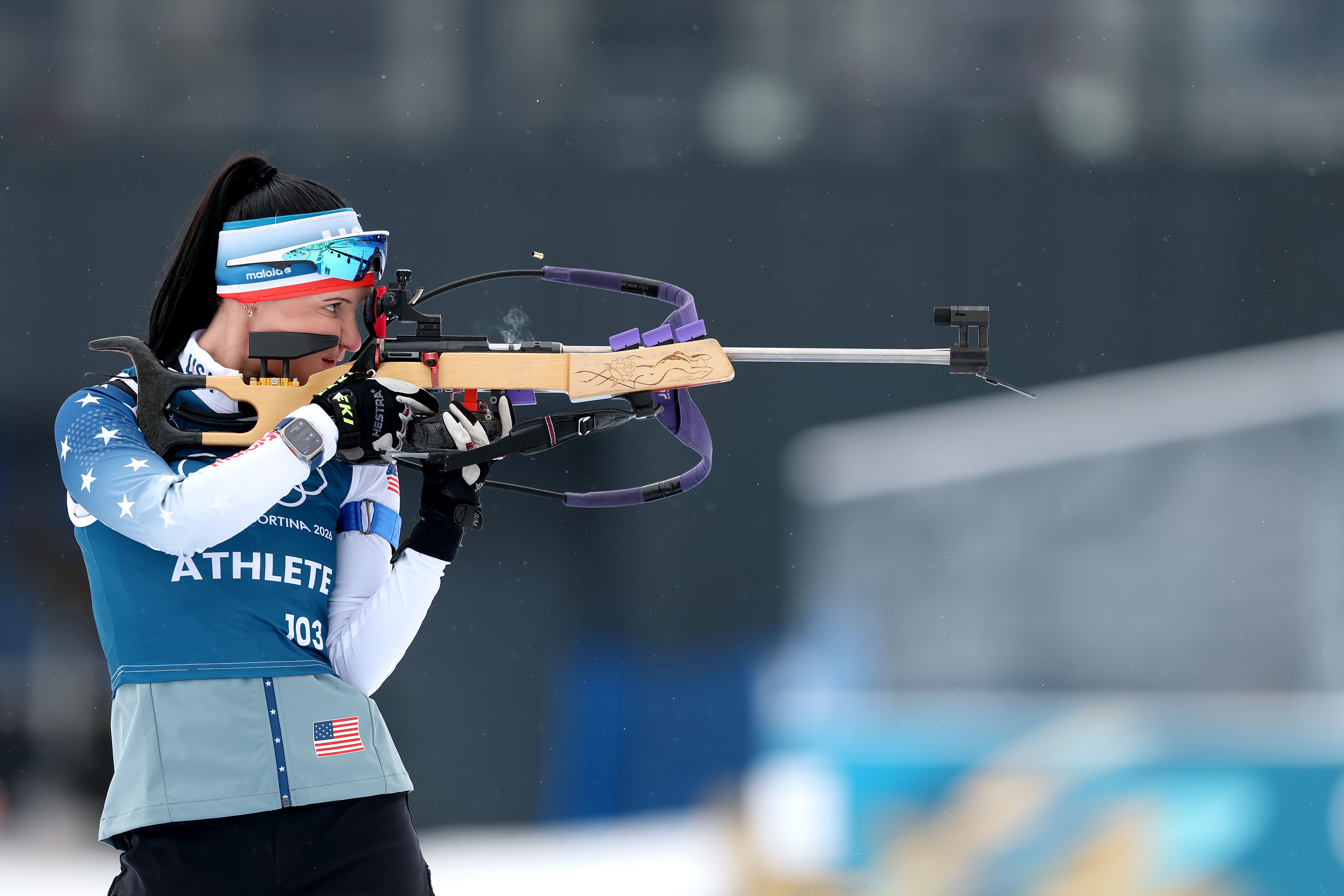 Female biathlete in blue and white USA uniform aiming a rifle, with visible breath vapor against a blurred snowy background, during a competitive event.