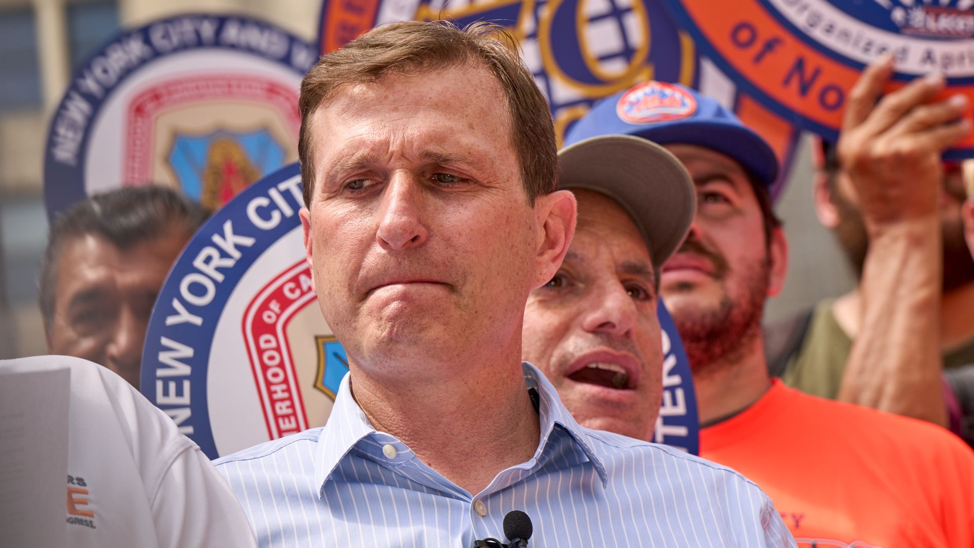 Close-up of a man in a light blue striped shirt with two lapel microphones at a rally; behind him are large circular banners in blue, orange, and red and a cheering crowd.