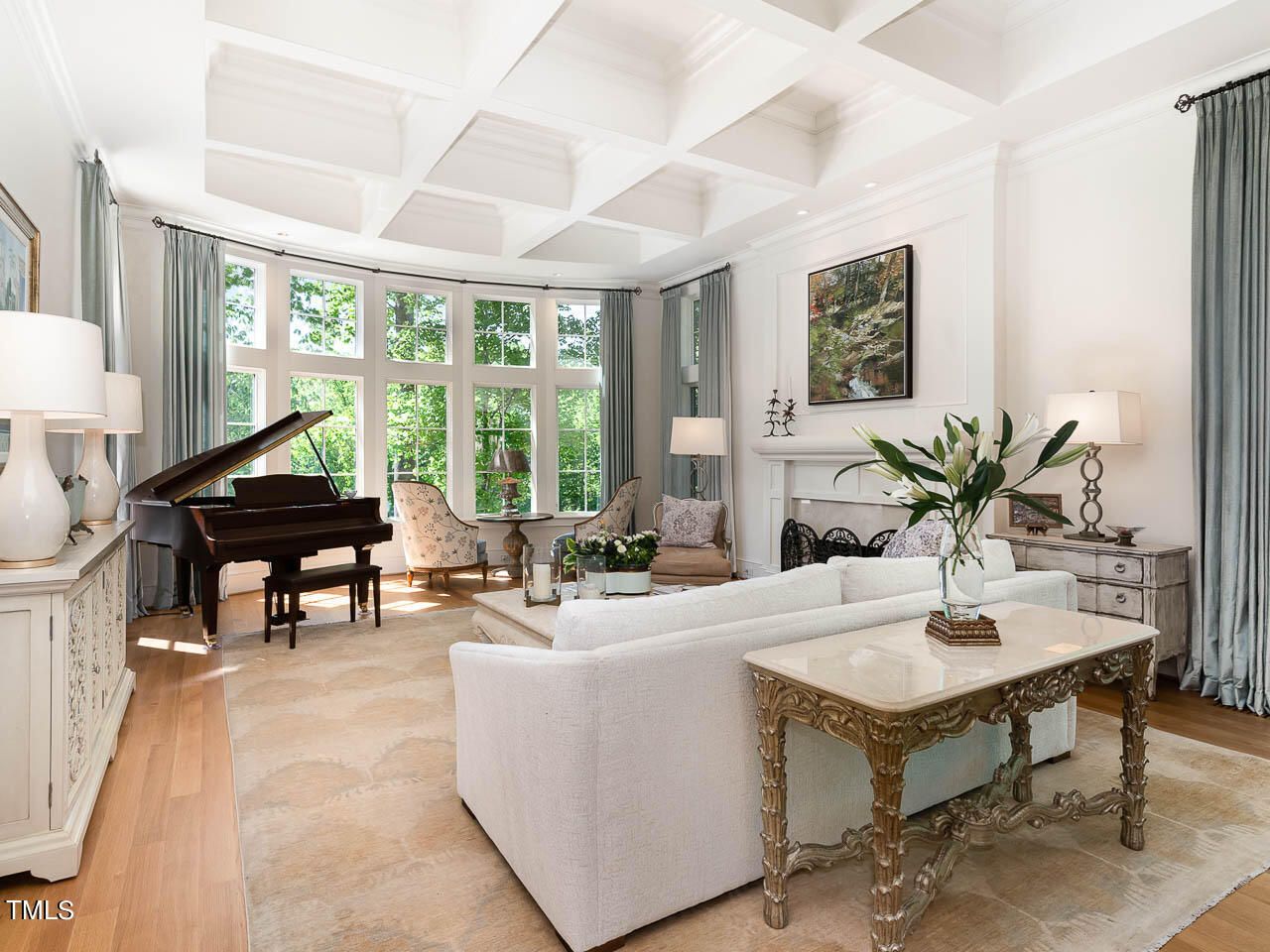 Bright living room with white walls, large windows with blue curtains, white sofa, ornate wooden table, grand piano, two armchairs, fireplace with a painting, and decorative lamps.