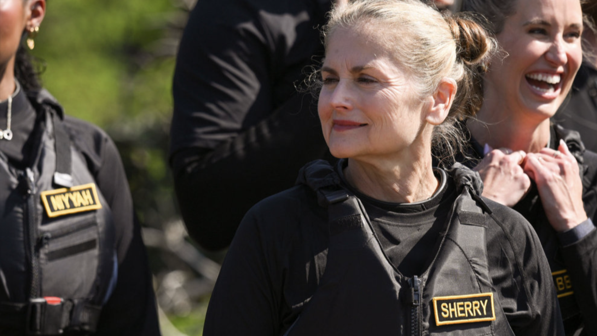 Three women in black vests outdoors. The center woman with light hair smiles slightly; the right woman laughs. A name patch on the center vest reads "SHERRY".