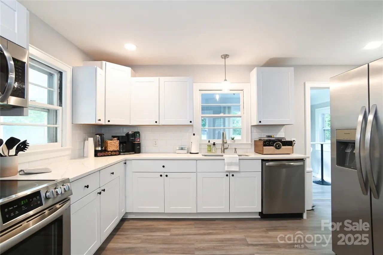 Bright modern kitchen with white cabinets, stainless steel appliances including oven, microwave, dishwasher, and refrigerator. Wood-style flooring and a window above the sink with hanging light.