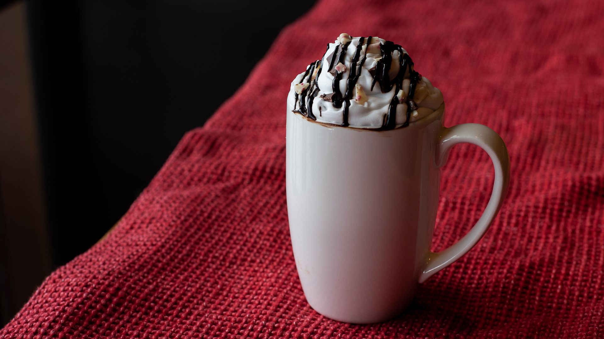 A mocha drink with whipped cream, chocolate drizzle and bits of peppermint bark in a white mug on a table.