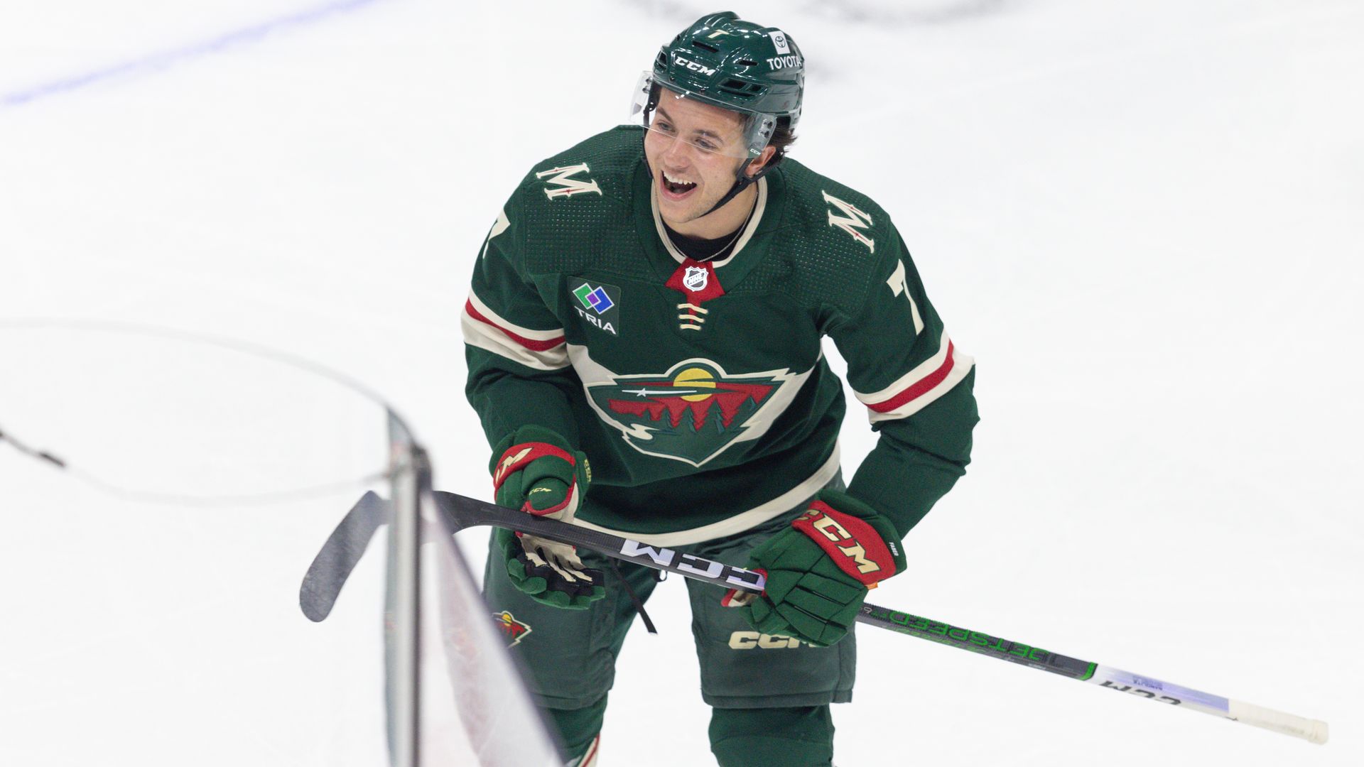 A hockey player in a forest green jersey smiles and holds his stick at waist level as he skates on a white sheet of ice