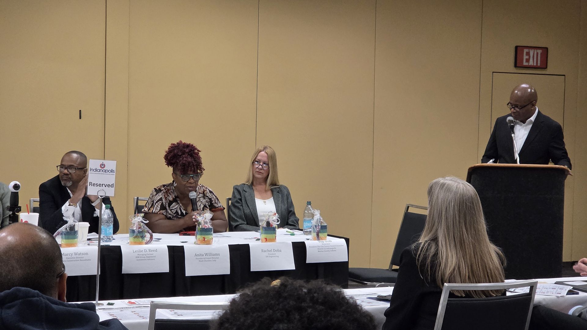 Panel discussion with four seated panelists and one standing speaker at a podium in a beige room, with audience members in the foreground.