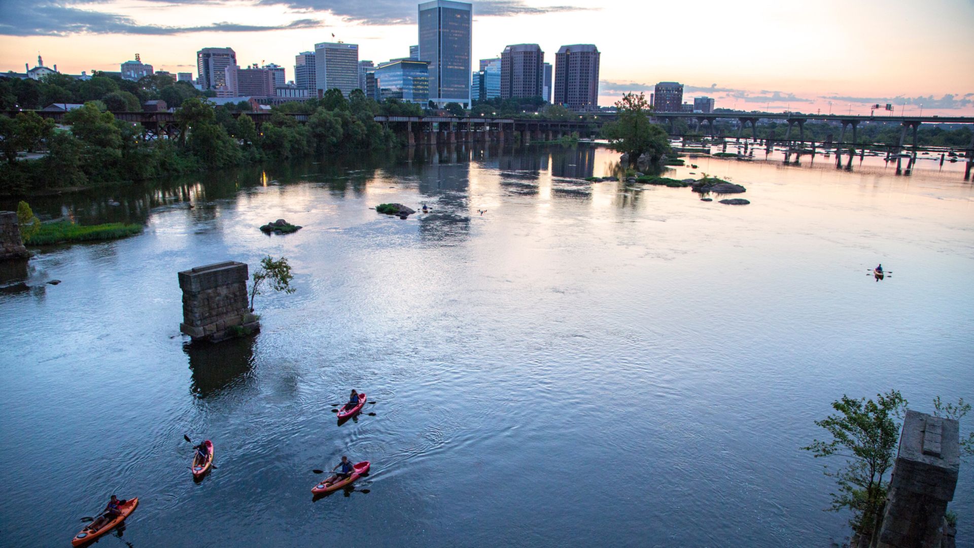 Five people kayaking in red kayaks on a calm river with a city skyline and bridges at sunset under a partly cloudy sky.