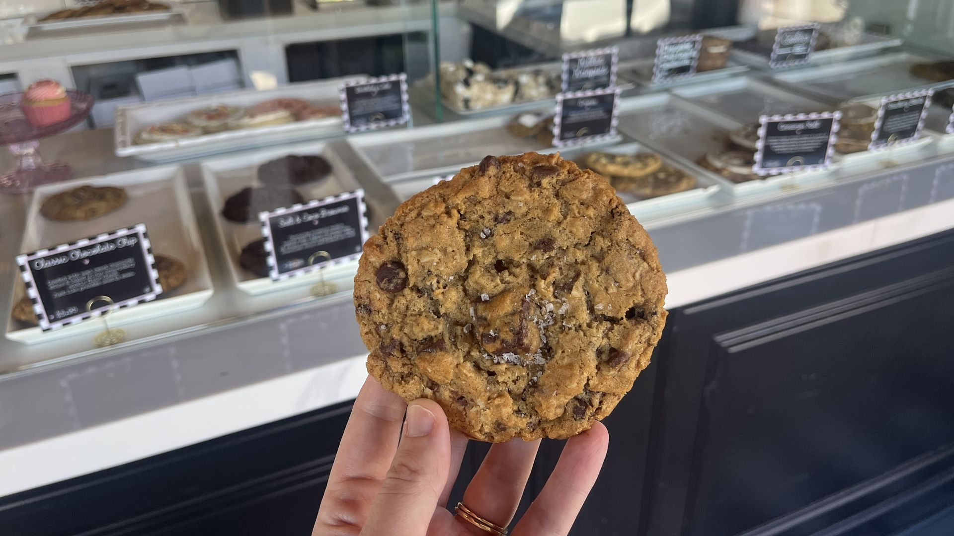 Close-up of a large, chunky oatmeal chocolate chip cookie with raisins held by a hand in front of a bakery display case filled with assorted cookies and pastries.