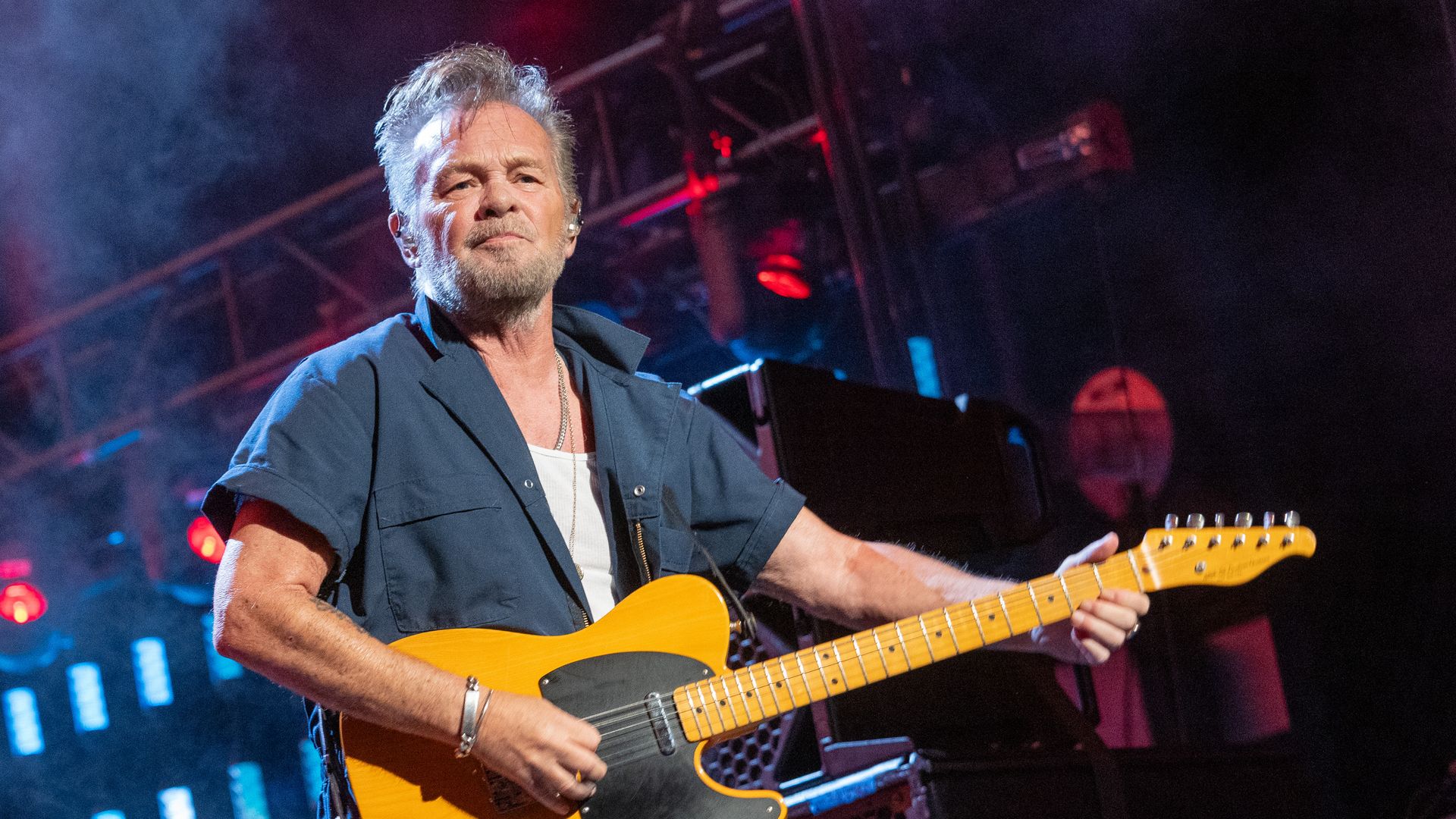 Man with gray hair and beard playing yellow electric guitar on stage wearing dark blue shirt over white undershirt, with red and blue stage lights and smoke in background.