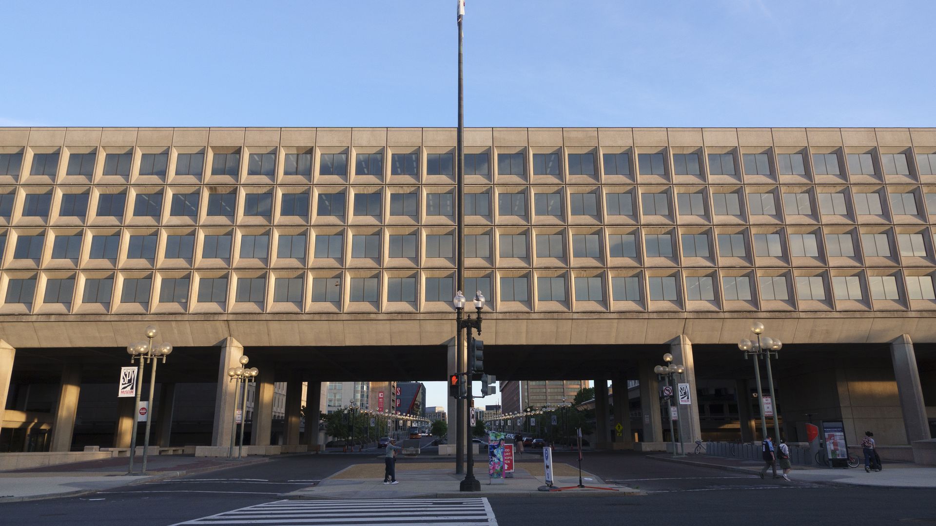 Wide shot of a beige office building with a central flagpole flying the American flag, set against a clear blue sky; street level with pedestrians and street lamps.