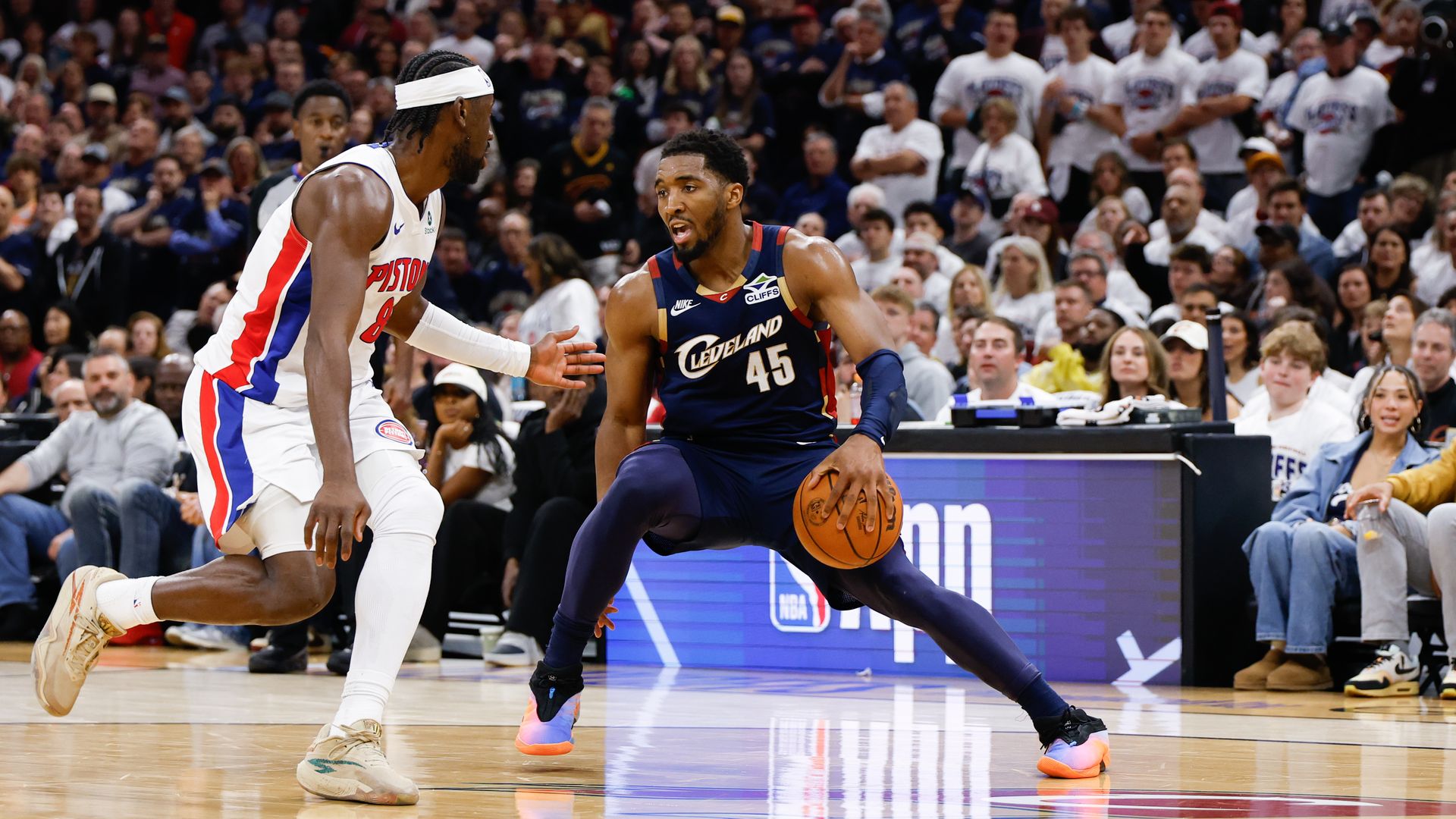 Donovan Mitchell (#45) in blue jersey crosses over PIstons player in white jersey on basketball court with crowd in background wearing white tees.