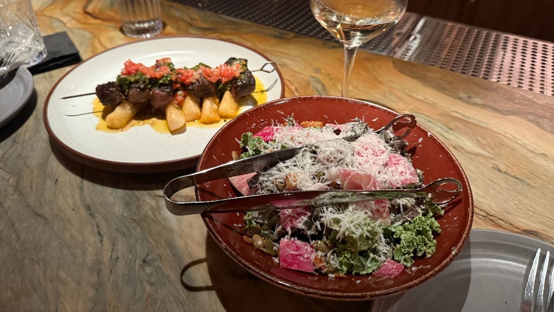 Dinner setting with a skewer of grilled meat and vegetables on fries, a leafy green salad topped with grated cheese and pink radish, a glass of white wine, and a glass of water on a marble countertop.