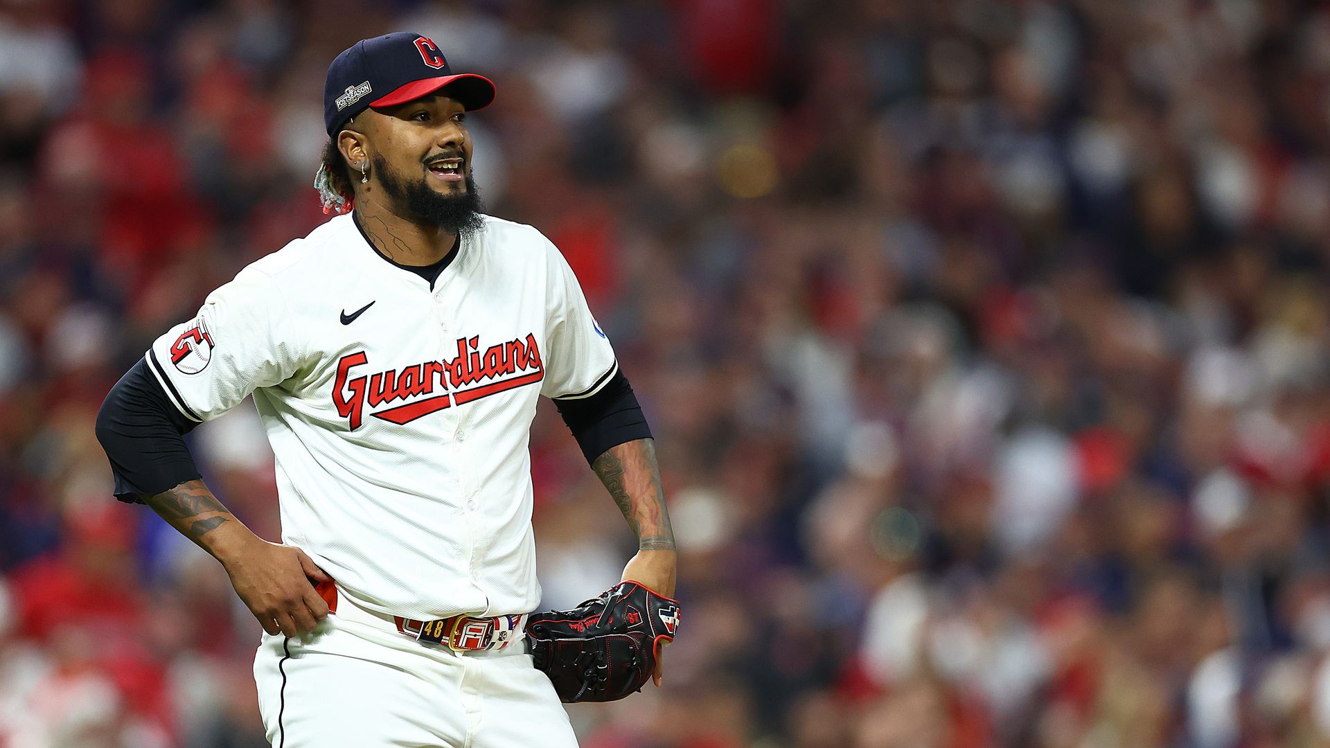 Baseball player in white "Guardians" uniform with black red hat and glove stands on field with crowd blurred in background.