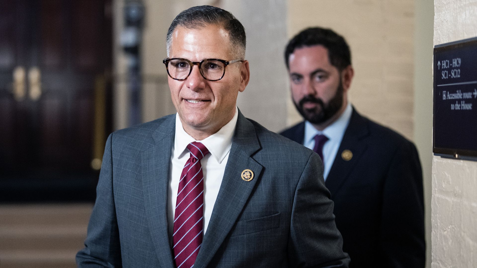 Rep. Marc Molinaro, wearing glasses and a gray suit, walking through a white brick hallway.