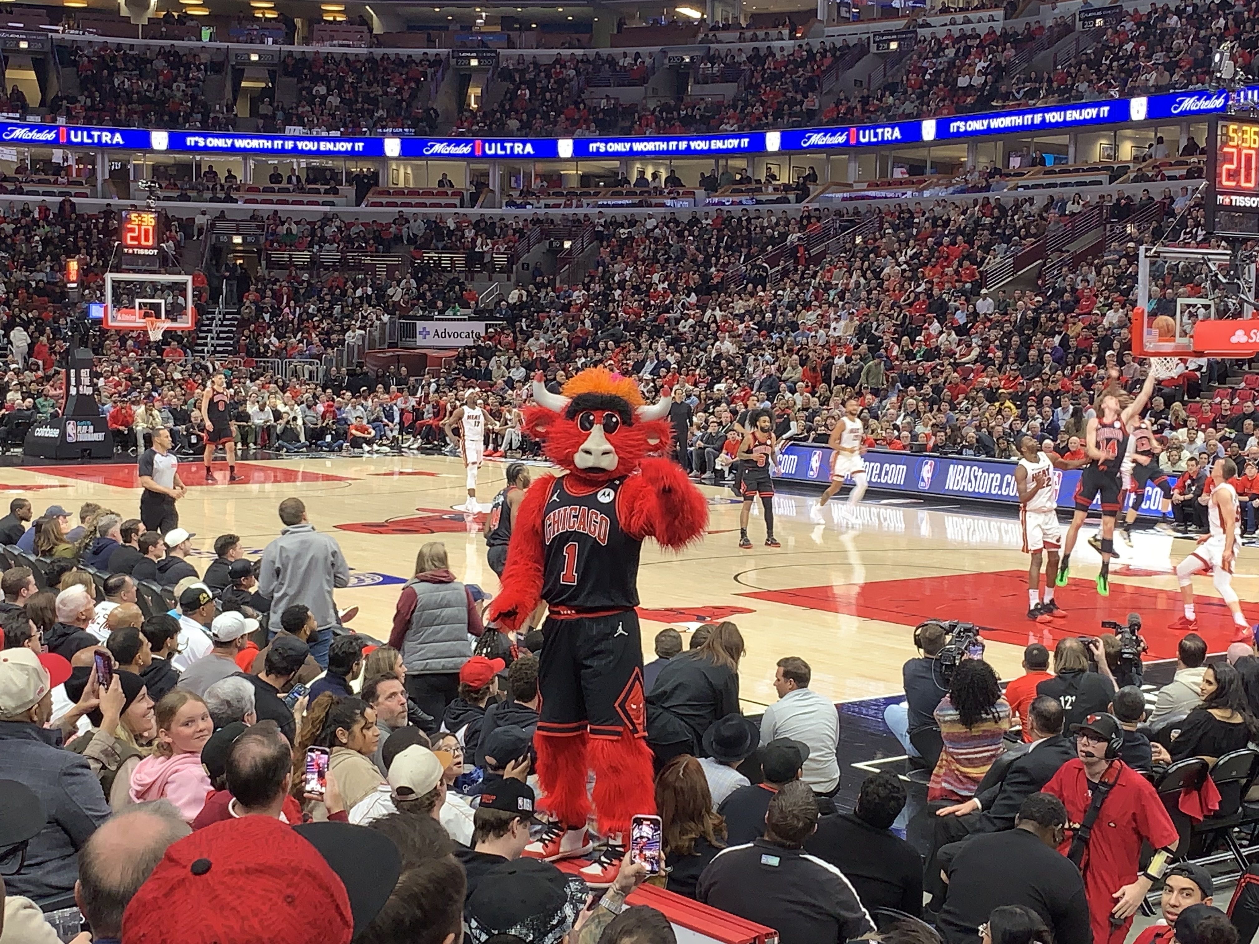 Busy basketball arena with a red Chicago Bulls mascot in a black jersey number 1 on court, surrounded by players, photographers, and a packed crowd of spectators.