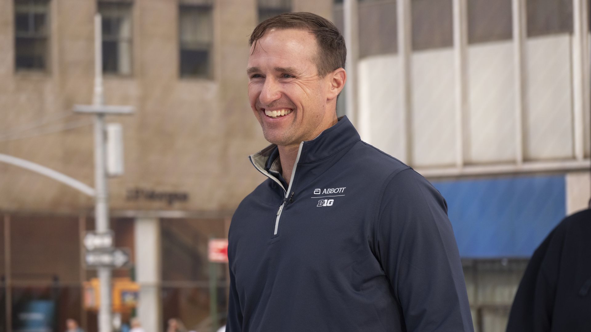 Smiling man wearing a navy blue zip-up jacket with "Abbott" and "Big Ten" logos, standing outdoors in an urban setting with blurred buildings and people in the background.
