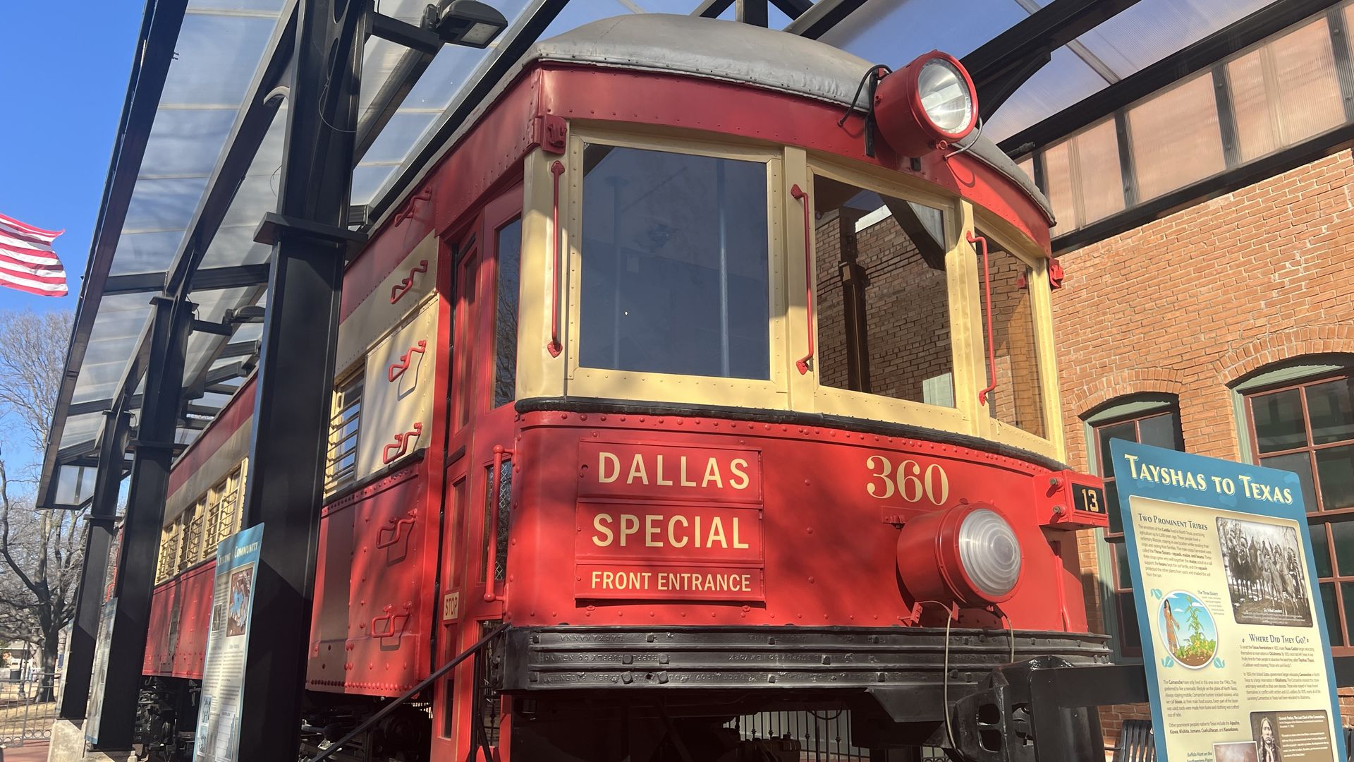 Red vintage trolley with gold trim and a large headlamp reads "Dallas Special Front Entrance" on a track under a glass canopy, beside a brick building and an info sign, with blue sky.
