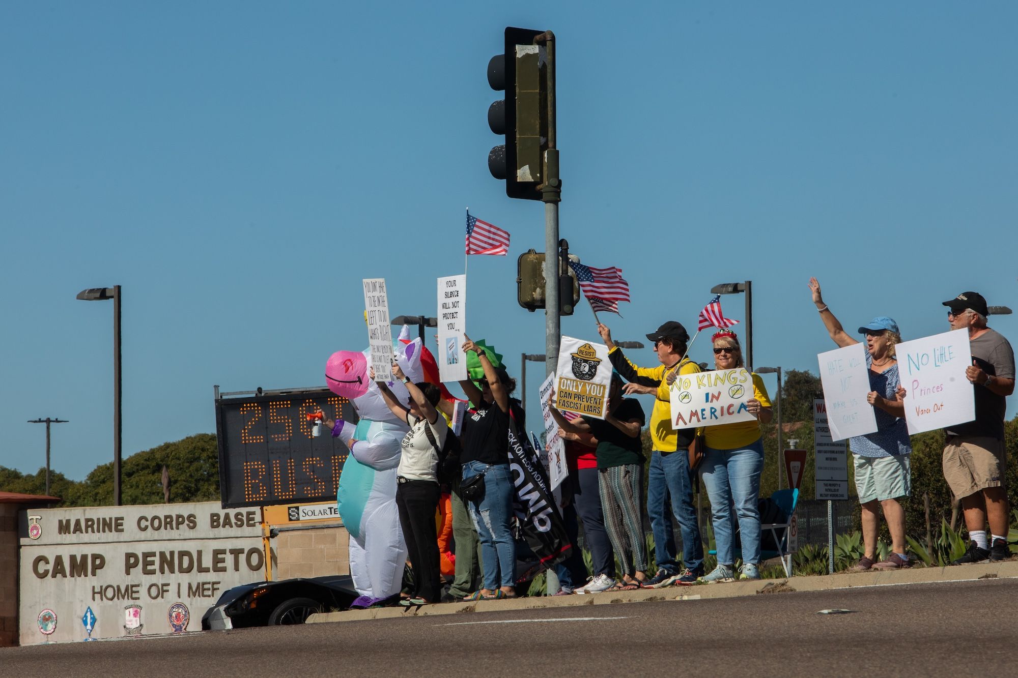 About a dozen protesters, including one wearing an inflatable unicorn costume, stand outside an entrance of Camp Pendleton Marine Corps Base waving flags and holding signs saying "No Kings in America."