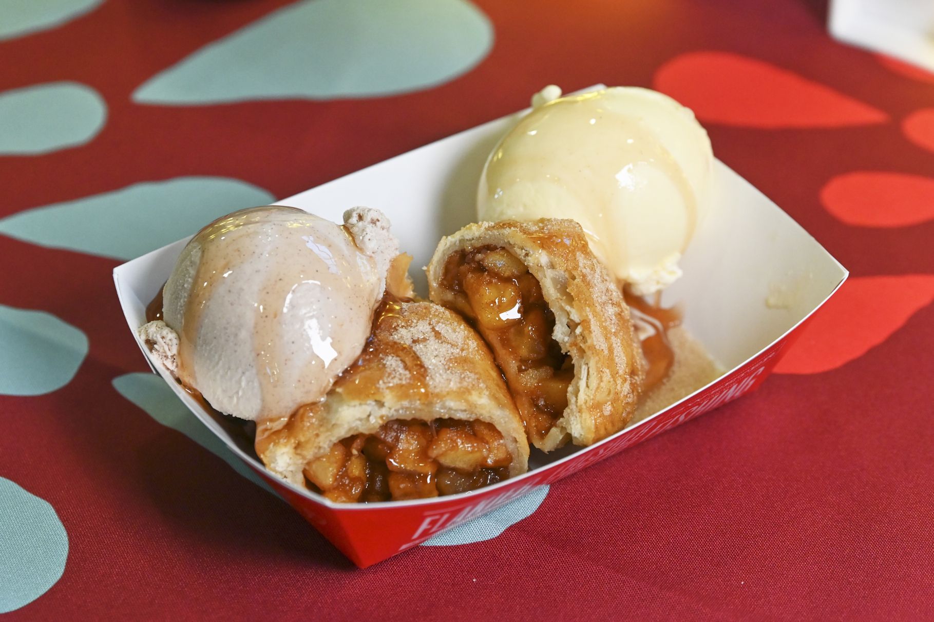 Two scoops of ice cream, one light brown and one pale vanilla, sit in a white tray beside a cut-open apple-filled pastry with caramel sauce, on a red table with teal shapes in the background.
