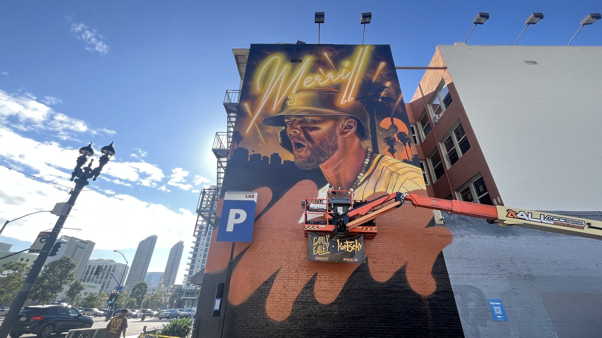 Large mural of Padres player Jackson Merrill in a batting helmet and striped jersey, with the word "Merrill" glowing above. Orange and black background with city skyline and palm trees.