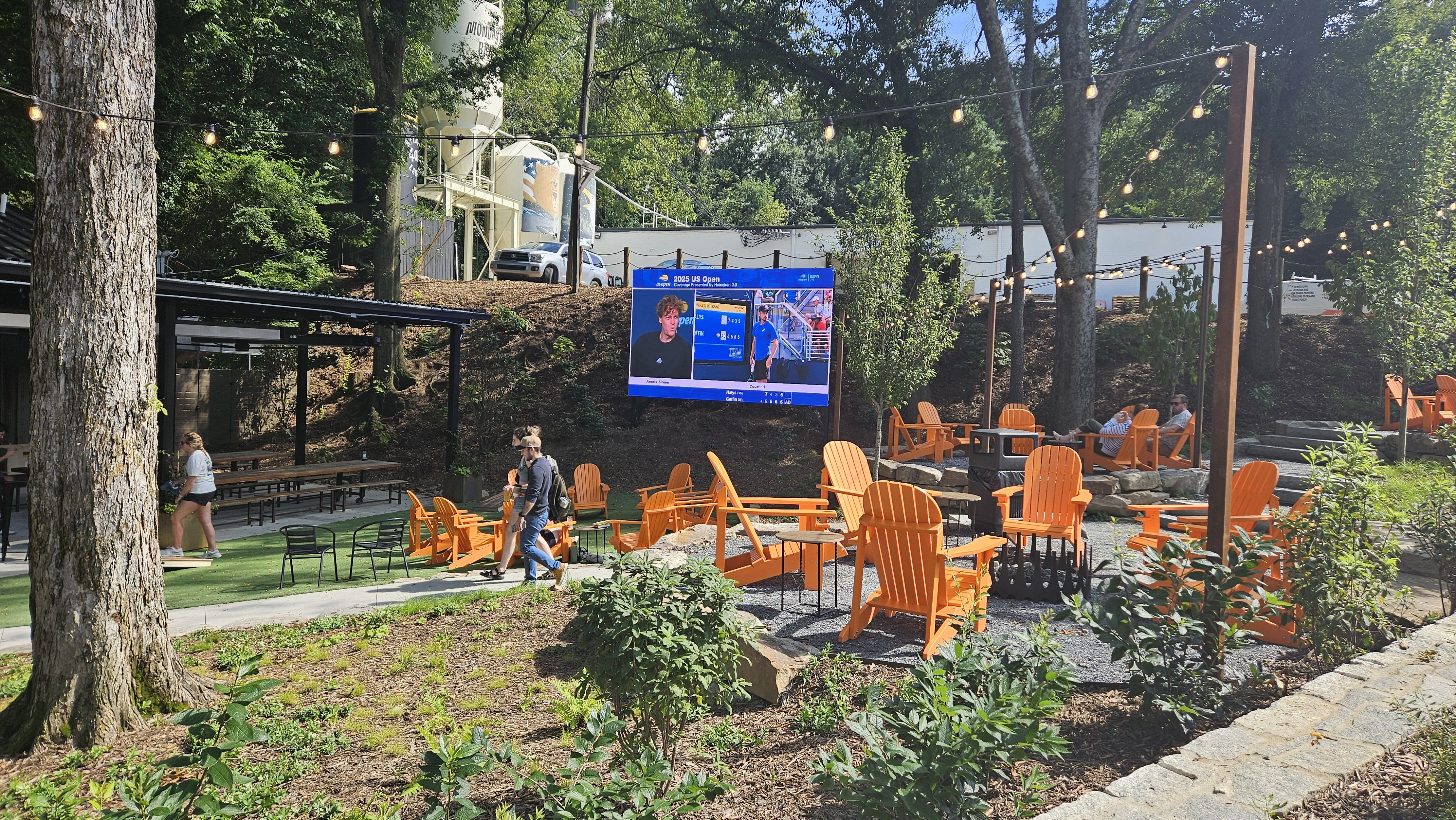 Outdoor seating area with bright orange Adirondack chairs around fire pits, string lights overhead, people walking and sitting, and a large screen showing 2025 US Open tennis coverage in a wooded setting.