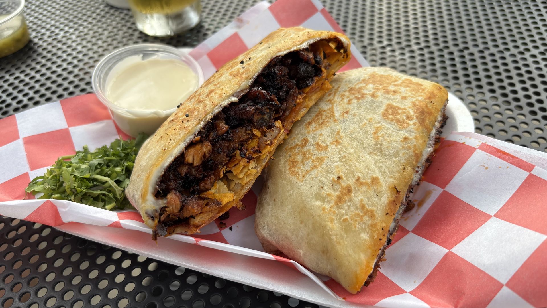 A crunchwrap cut in half with crispy browned pork al pastor on red-and-white checkered paper, served with a cup of white queso sauce and fresh green cilantro on a perforated metal table.