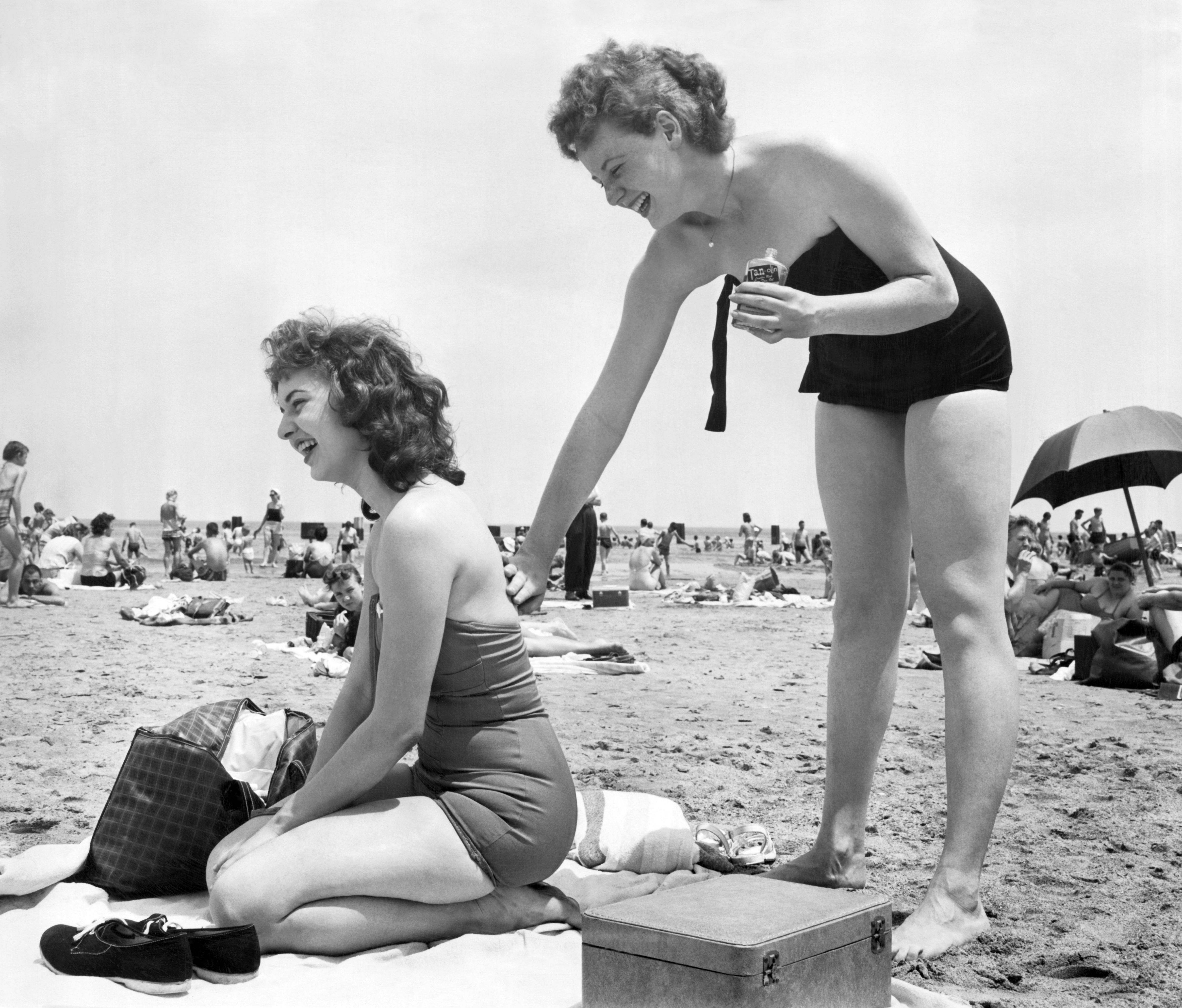 Black-and-white beach scene with many people. In foreground, a smiling woman in a one-piece sits on a towel; another woman in a black strapless swimsuit leans forward, holding a bottle.