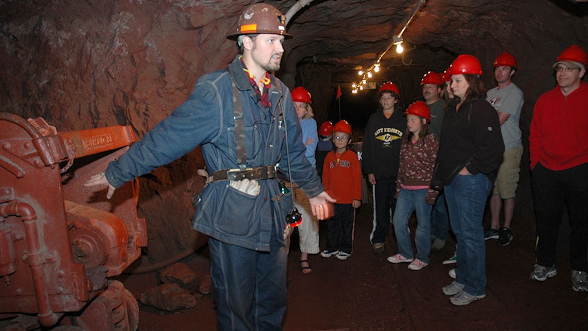 A tour guide in blue coveralls and a brown hard hat speaks to a group wearing red hard hats in a dim mine tunnel, with an orange mining machine on the left and string lights overhead.