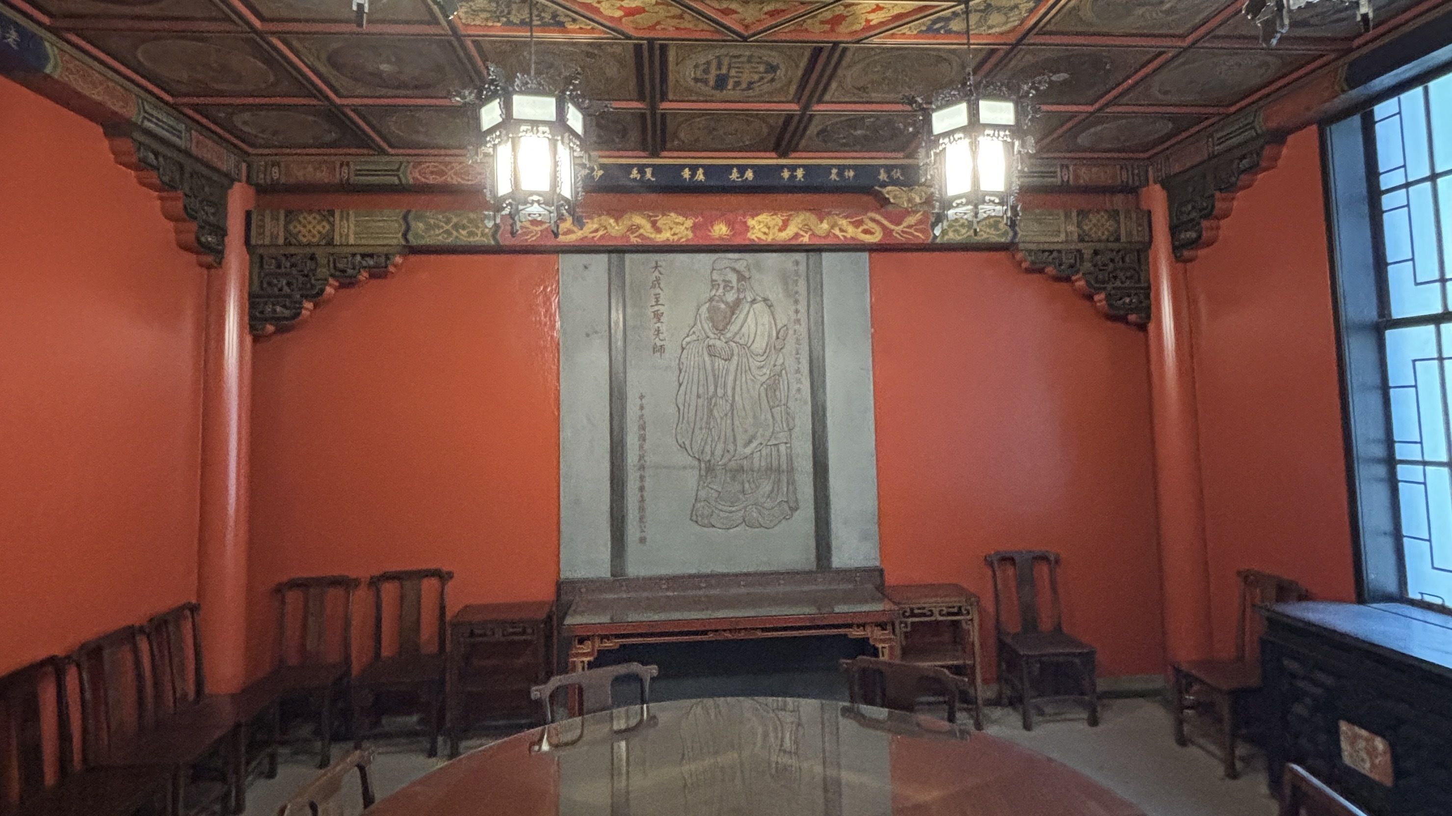 Red-walled Chinese-style room with an ornate carved ceiling and two hanging lanterns. A large scroll showing a bearded scholar hangs on the back wall, flanked by benches and a long glossy table, with blue windows to the right.
